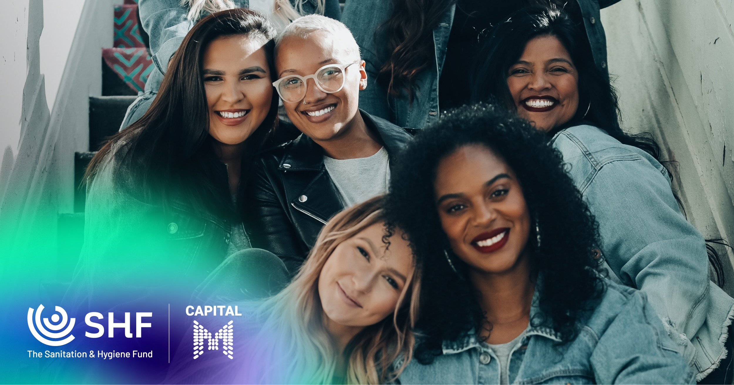 A diverse group of six women smiling and sitting on stairs, with some wearing denim jackets. The photo includes logos for The Sanitation & Hygiene Fund (SHF) and Capital M in the bottom left corner.