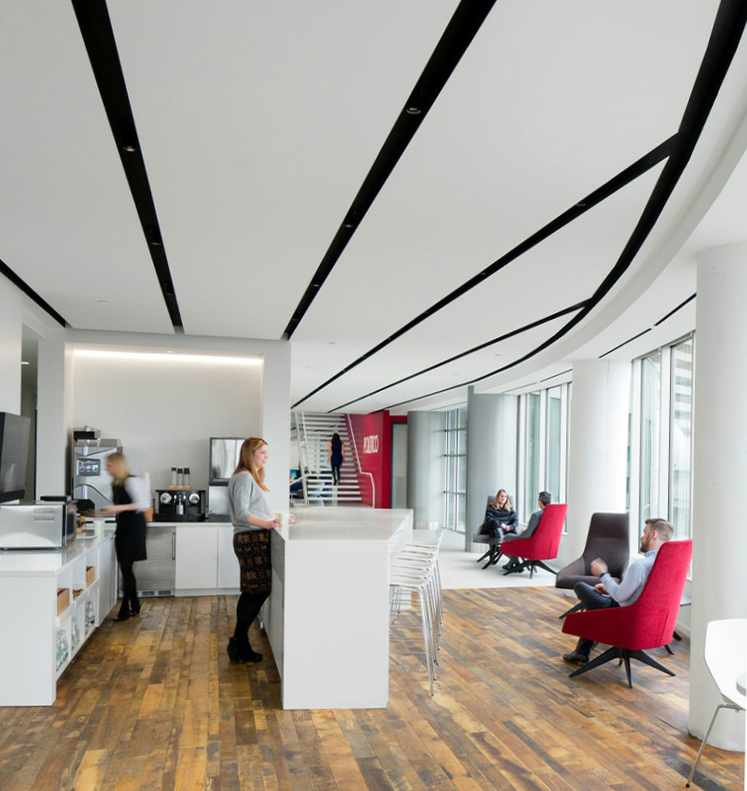 Modern office lounge area with white walls, large windows, and a wooden floor. There are chairs and people sitting, a woman at a white counter, and a coffee station in the background.