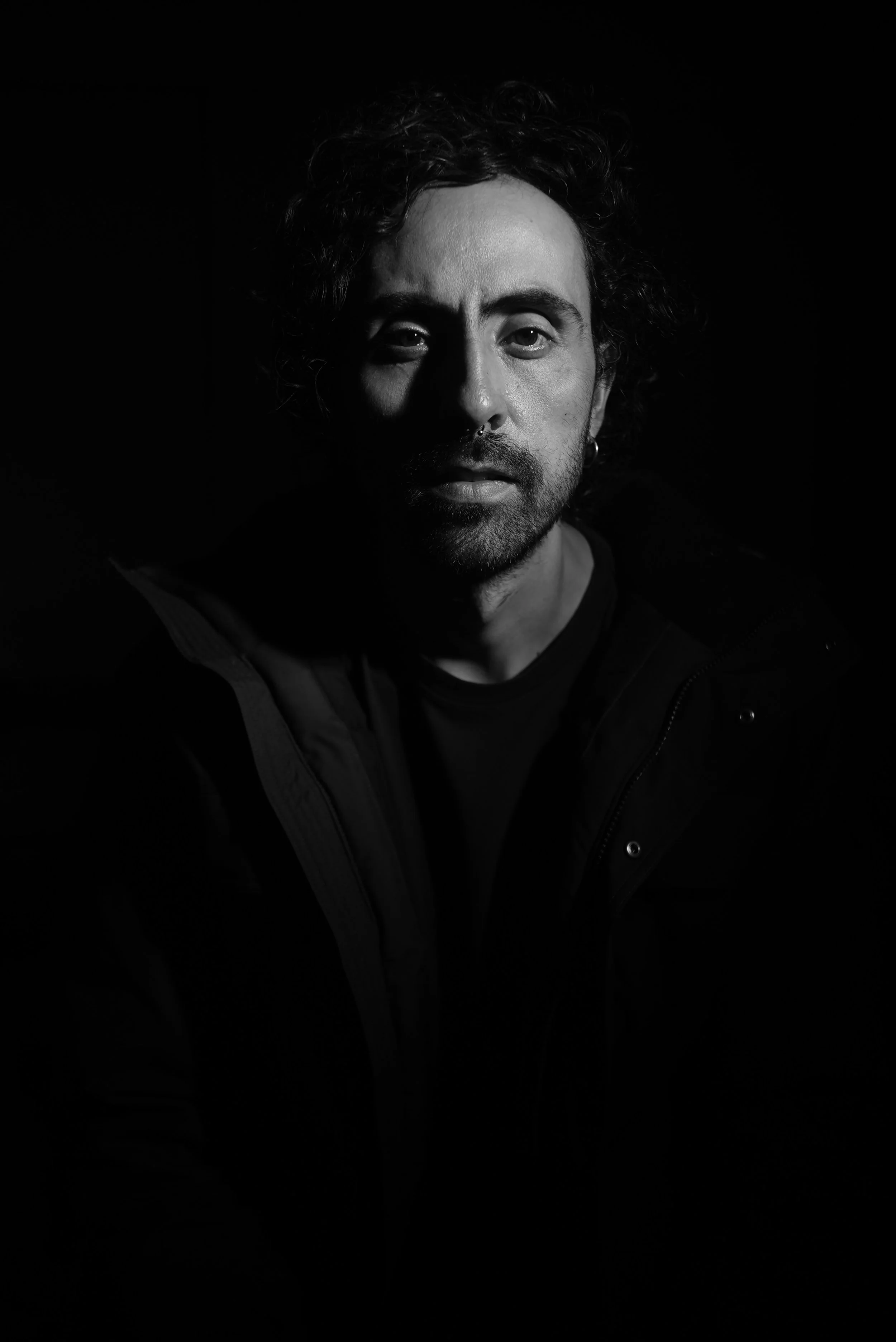 Black and white portrait of a man with curly hair, beard, and a septum piercing, wearing a dark jacket, with dramatic lighting on one side of his face.
