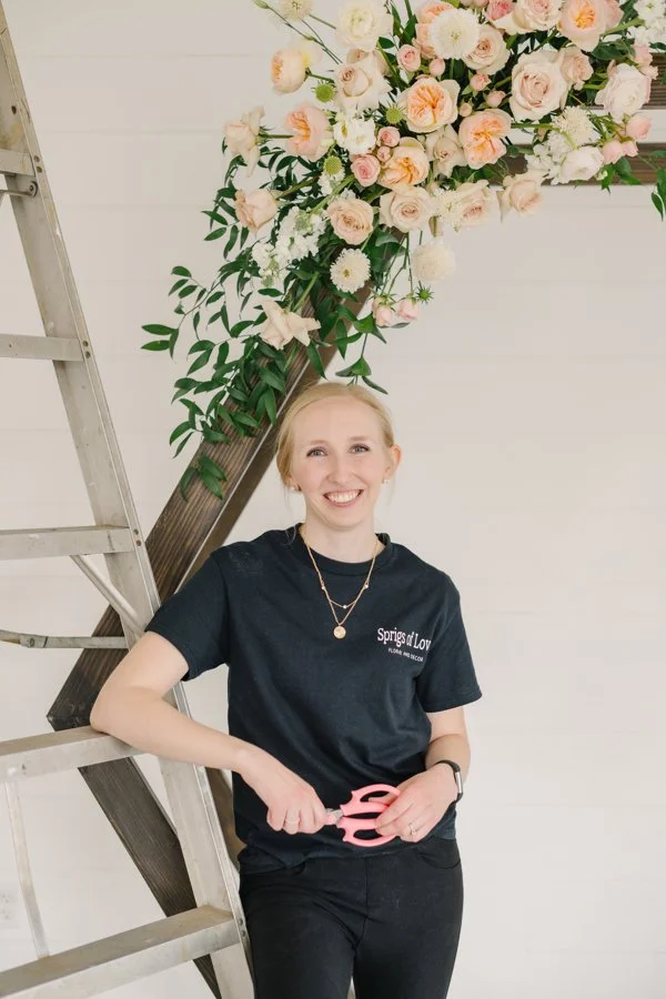 Utah wedding florist stands in front of a peach and pink ceremony arch and ladder.