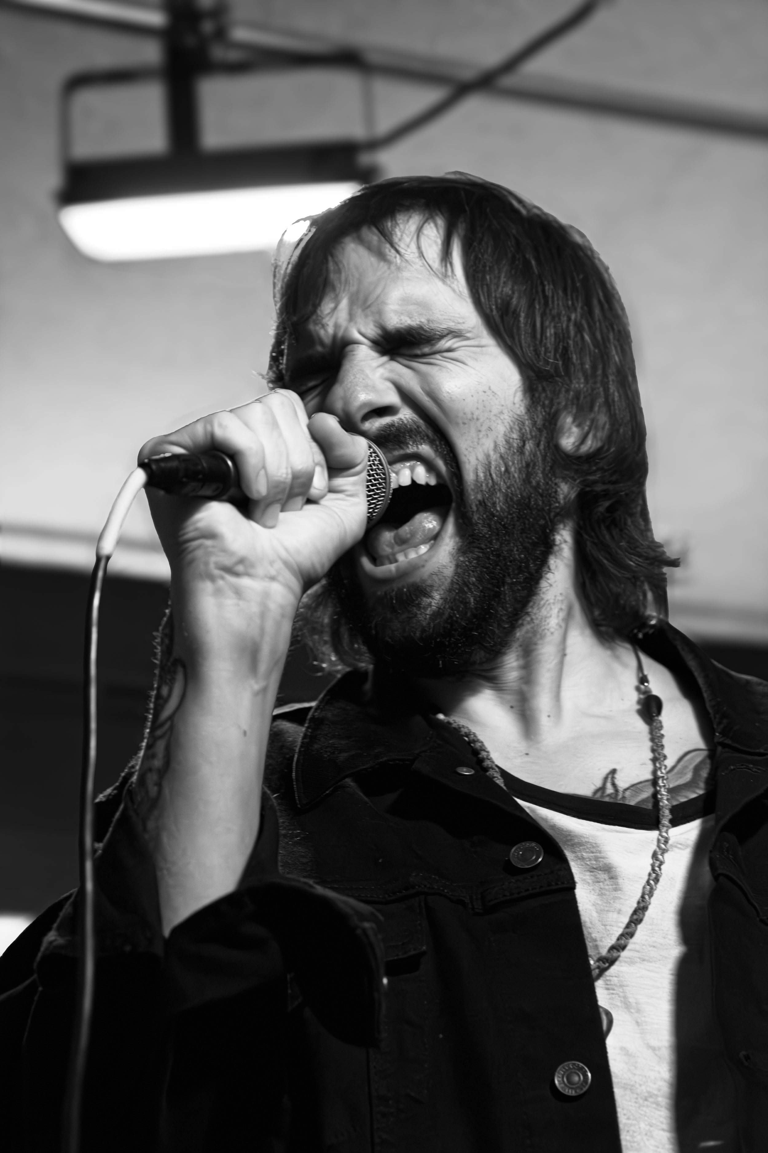 A Black and white photo of George Hamelin, the male lead singer of Manic Wave, singing passionately into a microphone, eyes closed, with long dark hair and a beard, wearing a dark jacket, a necklace, and a t-shirt.