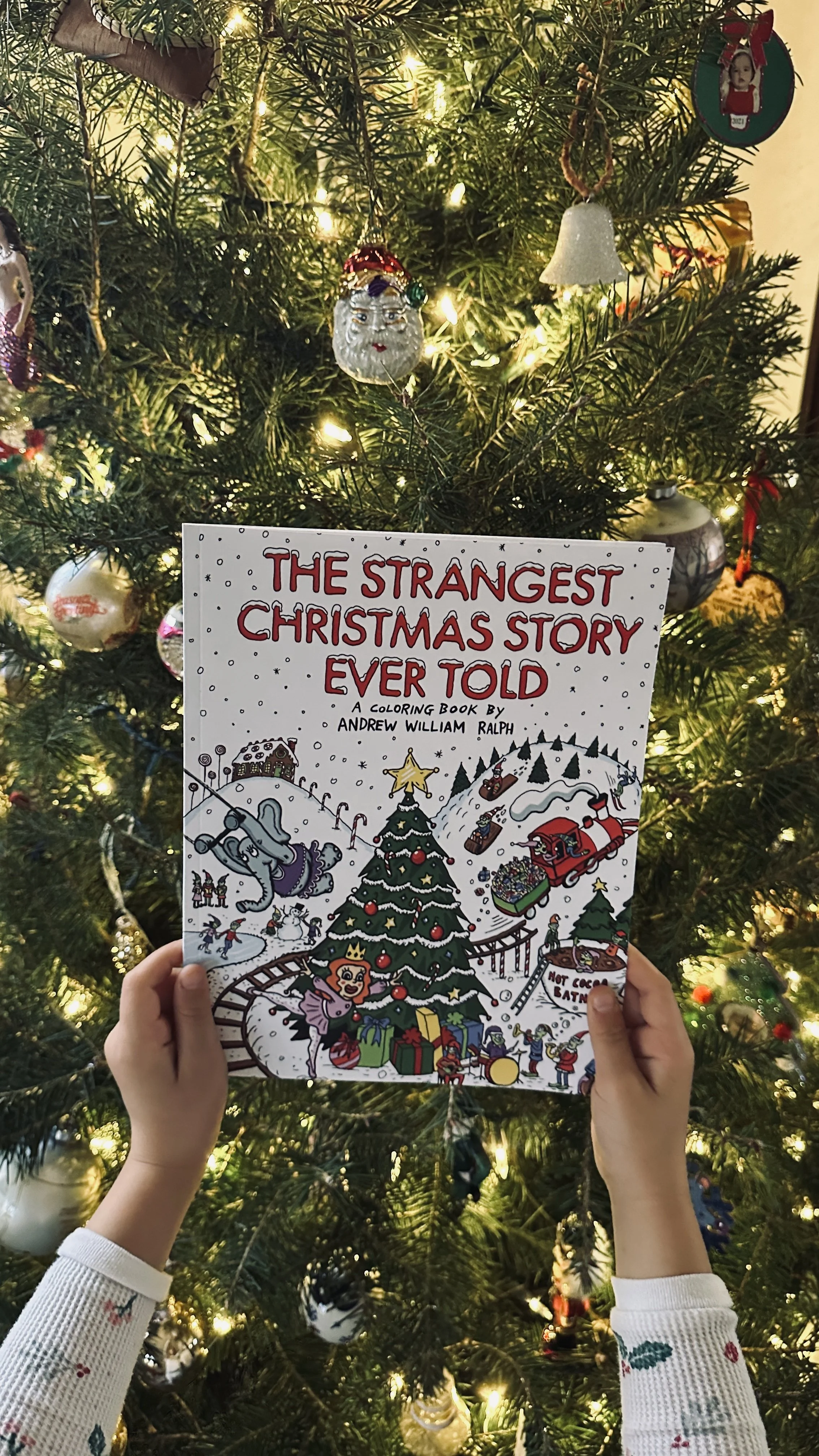 A person wearing festive Christmas sweater holding a coloring book titled 'The Strangest Christmas Story Ever Told' in front of a decorated Christmas tree with lights and ornaments.