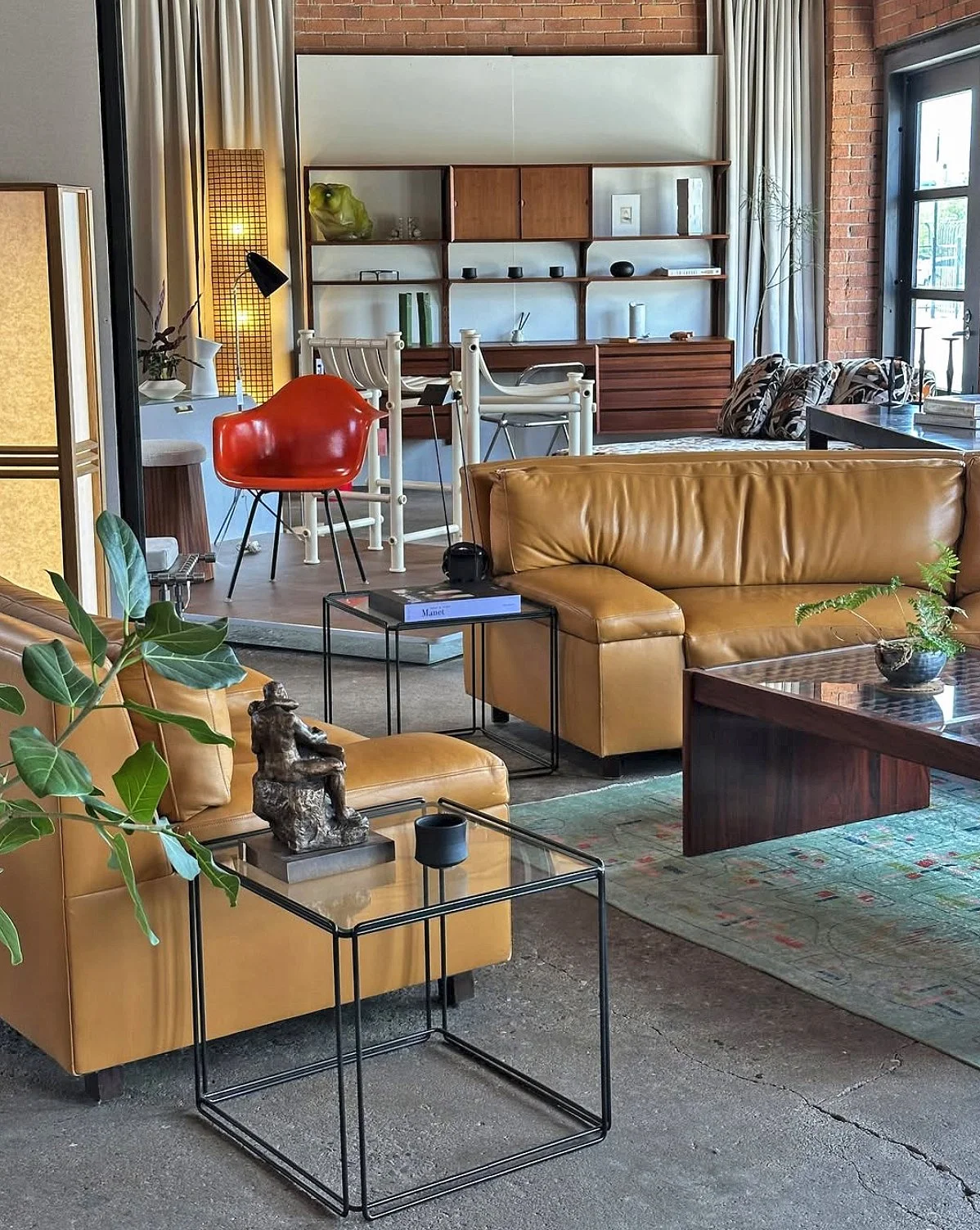 Saturday Generation showroom with stylish leather sofas, glass side tables, indoor plants, and a wooden bookshelf with decorative items. Background features a space with a Eames chair, danish shelving, and sculptures in a loft-like space.