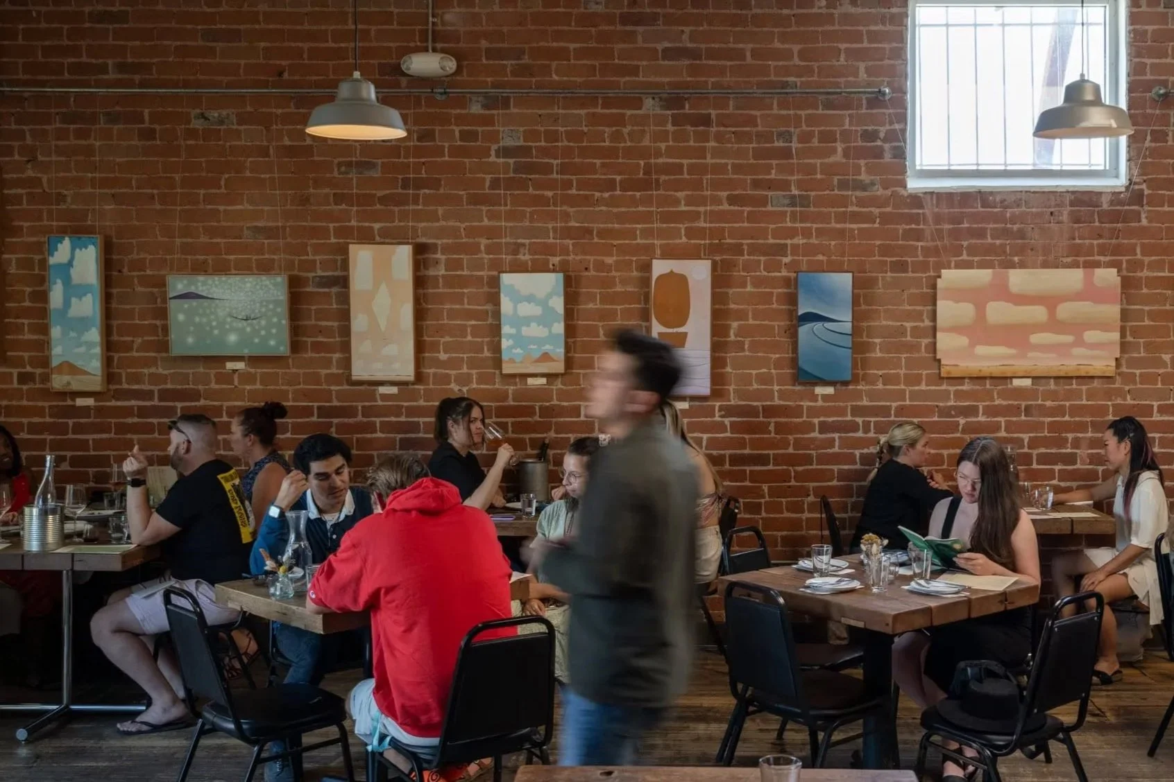 People dining at Five Points Market and Restaurant with a brick wall and paintings, some people are talking, one person reading a menu, and a blurred waiter walking by.