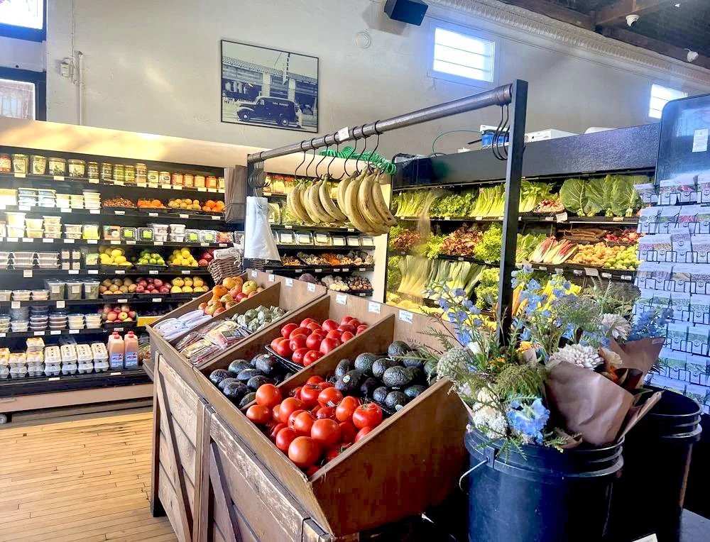 Fresh produce display at Time Market, including tomatoes, avocados, bananas, and flowers, with shelves of dairy products and leafy greens in the background.