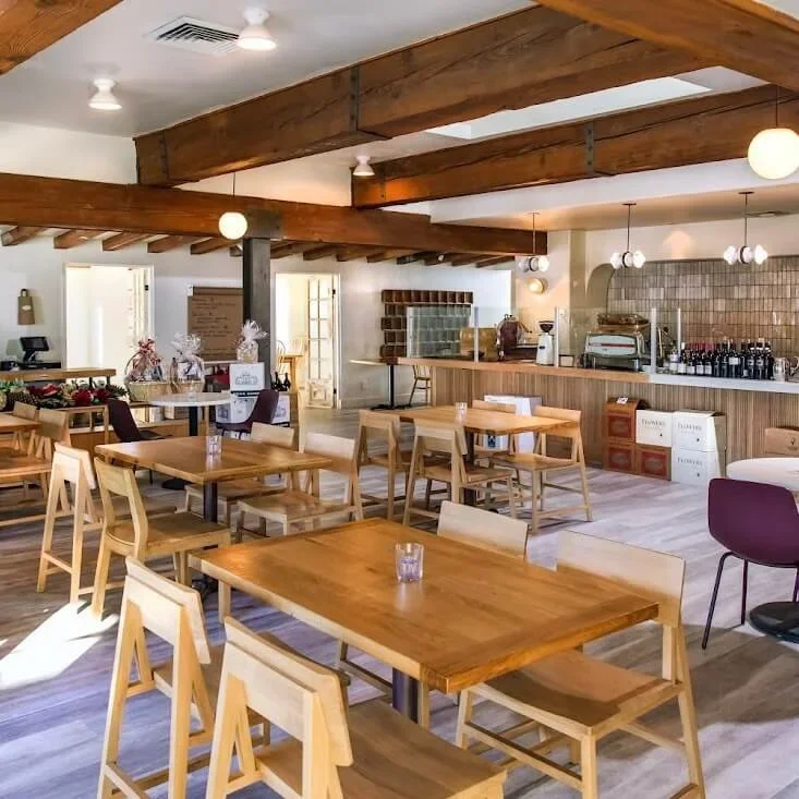 Locale cafe with wooden tables and chairs, soft ceiling lighting, and a bar area with glasses and bottles in the background.