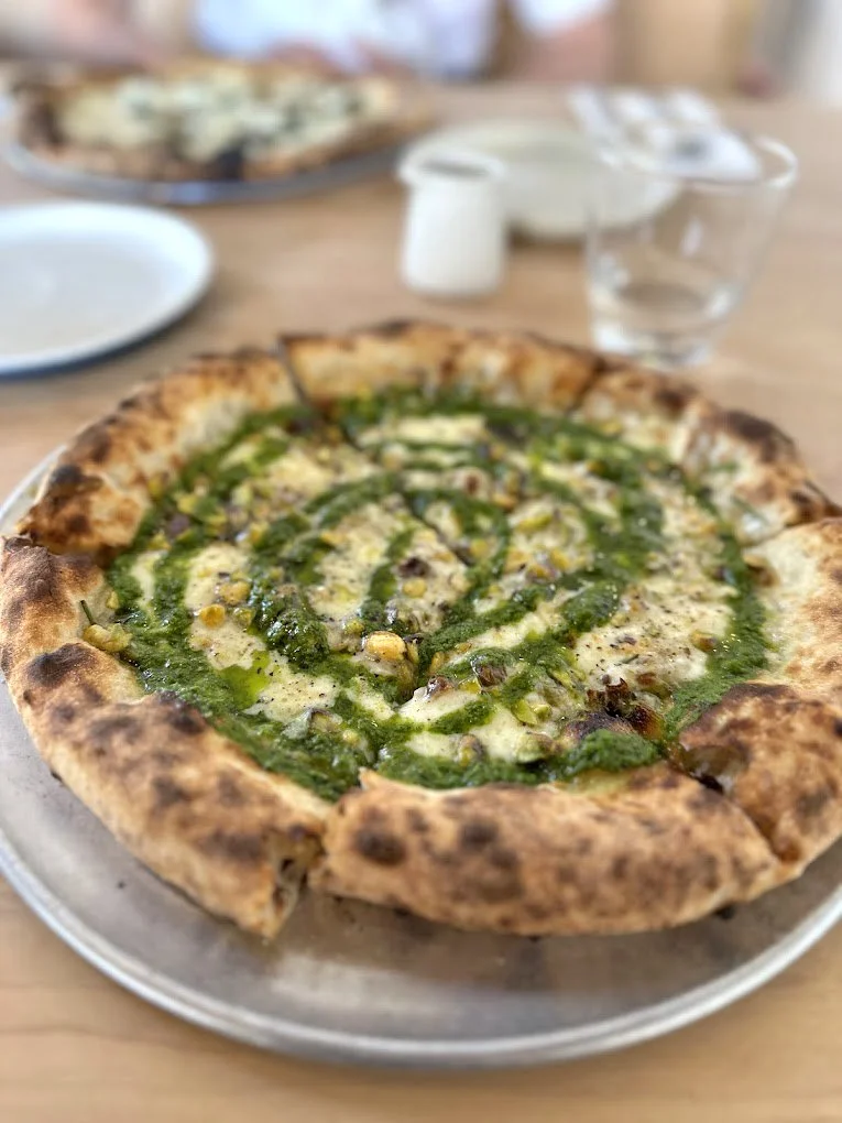 A Neapolitan pizza with green sauce, cheese, and toppings on a silver tray, with a slice missing, on a wooden table at Anello in Tucson.