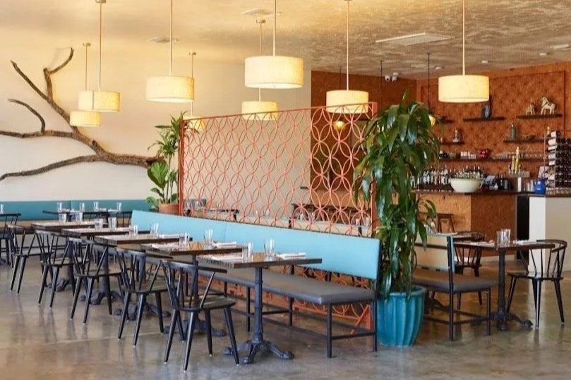 The interior of Tito & Pep with tables set for dining, hanging pendant lights, decorative wall art resembling tree branches, and a partition with a geometric pattern separating the dining area from the bar area.