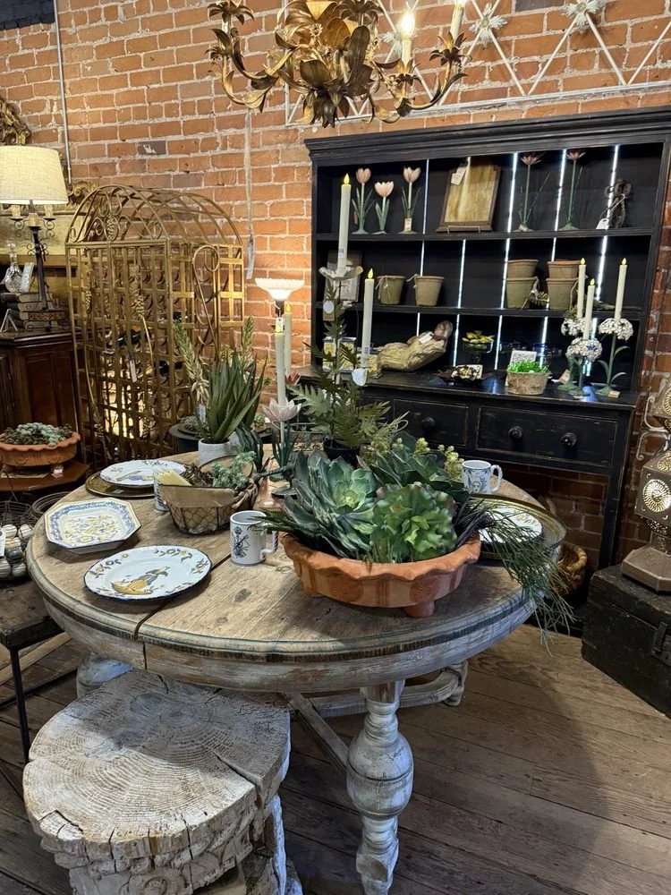 The showroom at Gather Vintage Market shows a rustic dining room with a round wooden table decorated with a large plant arrangement in a terracotta pot, vintage china plates, and mismatched mugs.