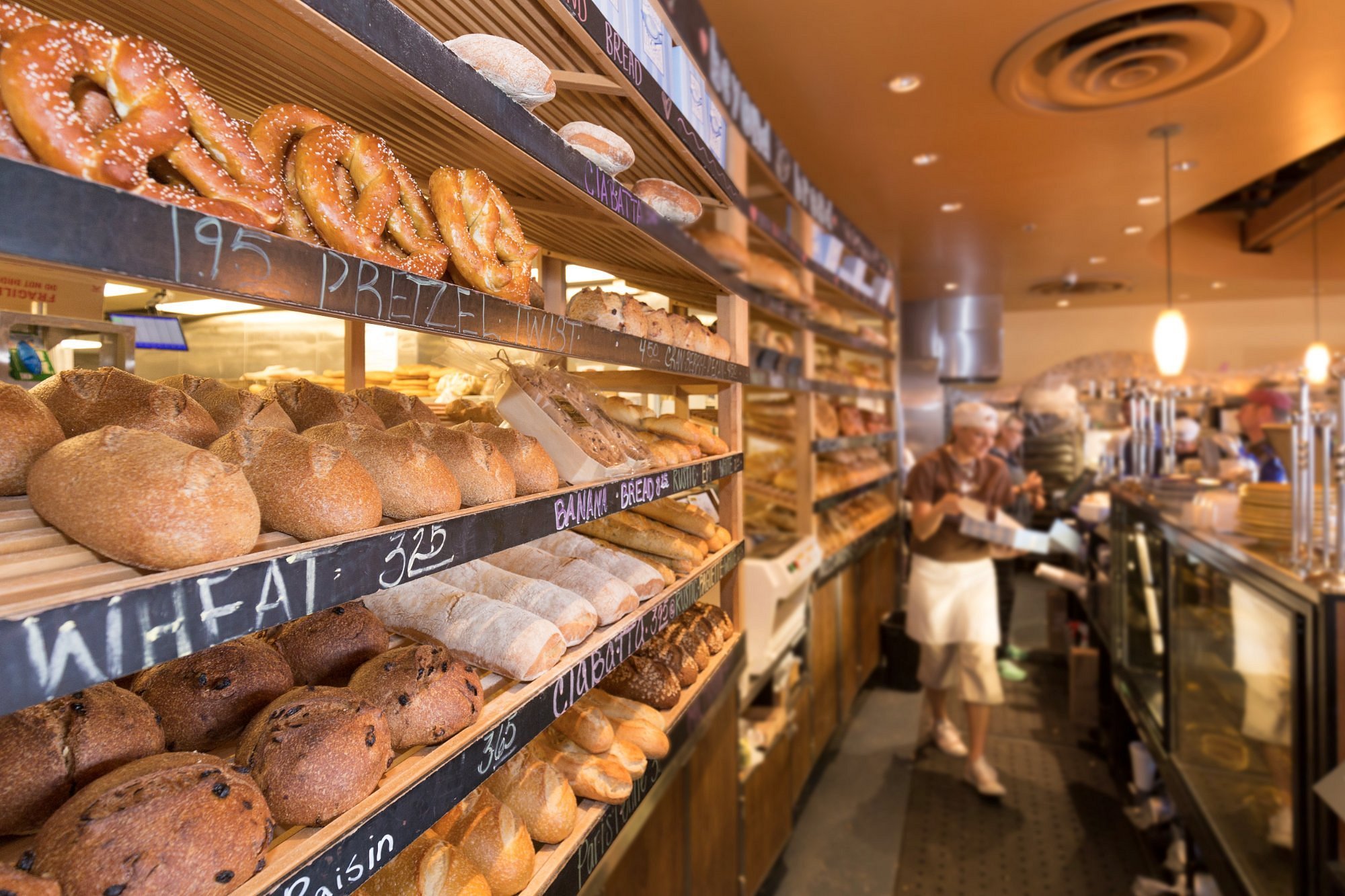 Selection of various bakery bread and baked goods displayed on wooden shelves in Beyond Bread restaurant, with customers and staff in the background.