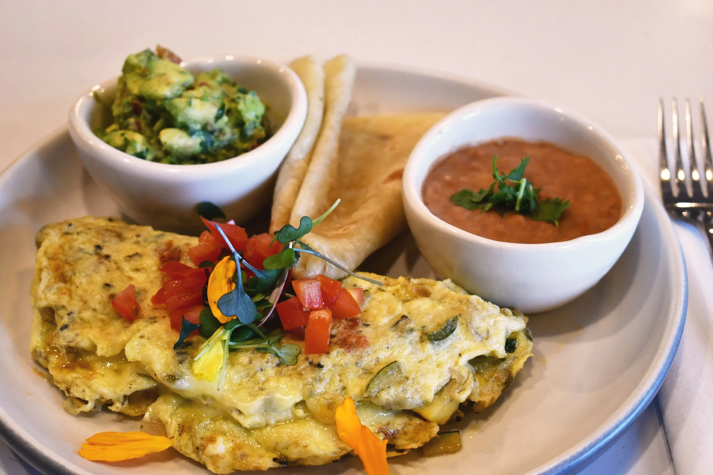 Le Buzz Caffe breakfast plate with omelette topped with diced tomatoes and microgreens, guacamole, red sauce with cilantro, and two breadsticks on a white plate.