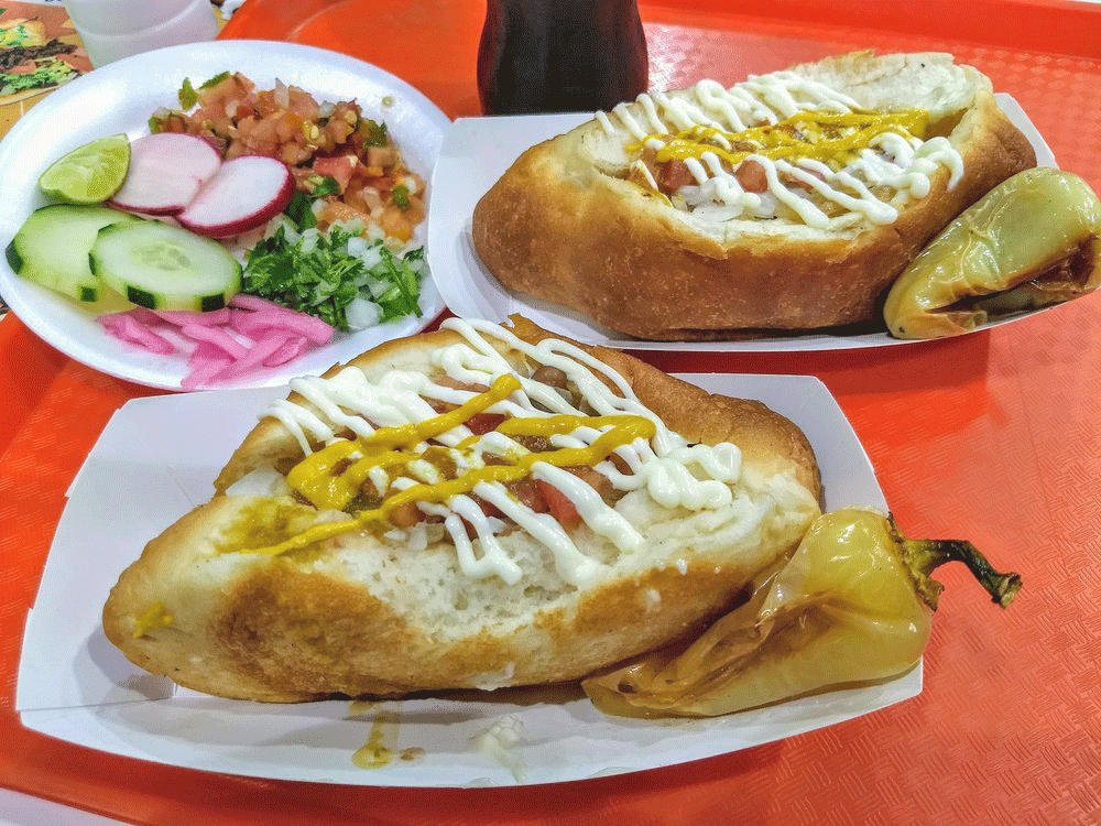 Two plates of Sonoran hotdogs with stuffed fresh-made buns topped with mustard and mayonnaise, served with grilled peppers at Guero Canelo in Tucson.