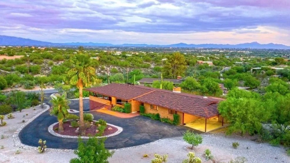 A single-story house with a red-tiled roof, surrounded by desert landscaping, palm trees, and a dark asphalt driveway, with a backdrop of a cityscape and mountains under a cloudy sky.