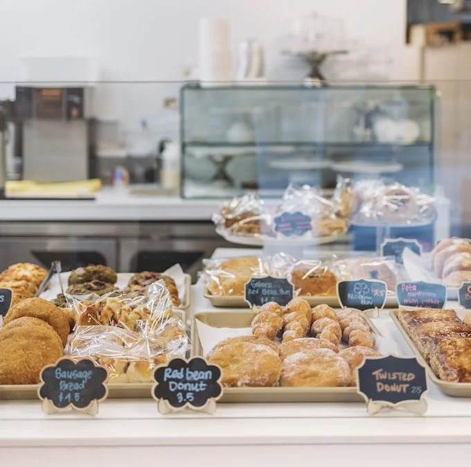 Assorted baked goods in a bakery display at HeeMee Bakery, including  red bean donuts, twisted donuts, and various pastries, with small chalkboard signs indicating their names and prices.