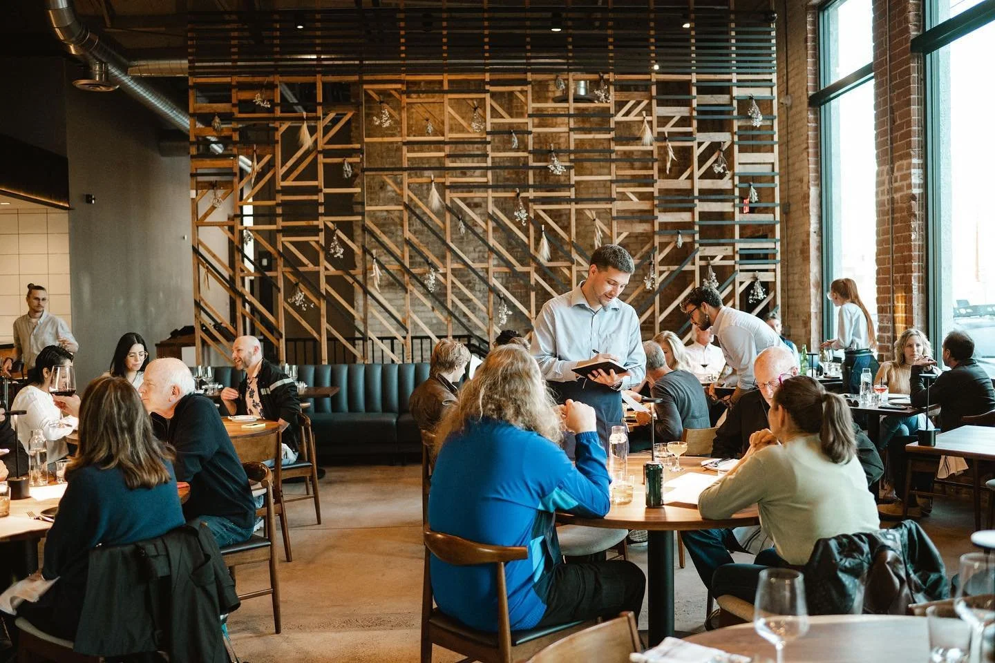 People dining at BATA restaurant with a modern brick and wood interior, large windows, and decorative wood wall art.