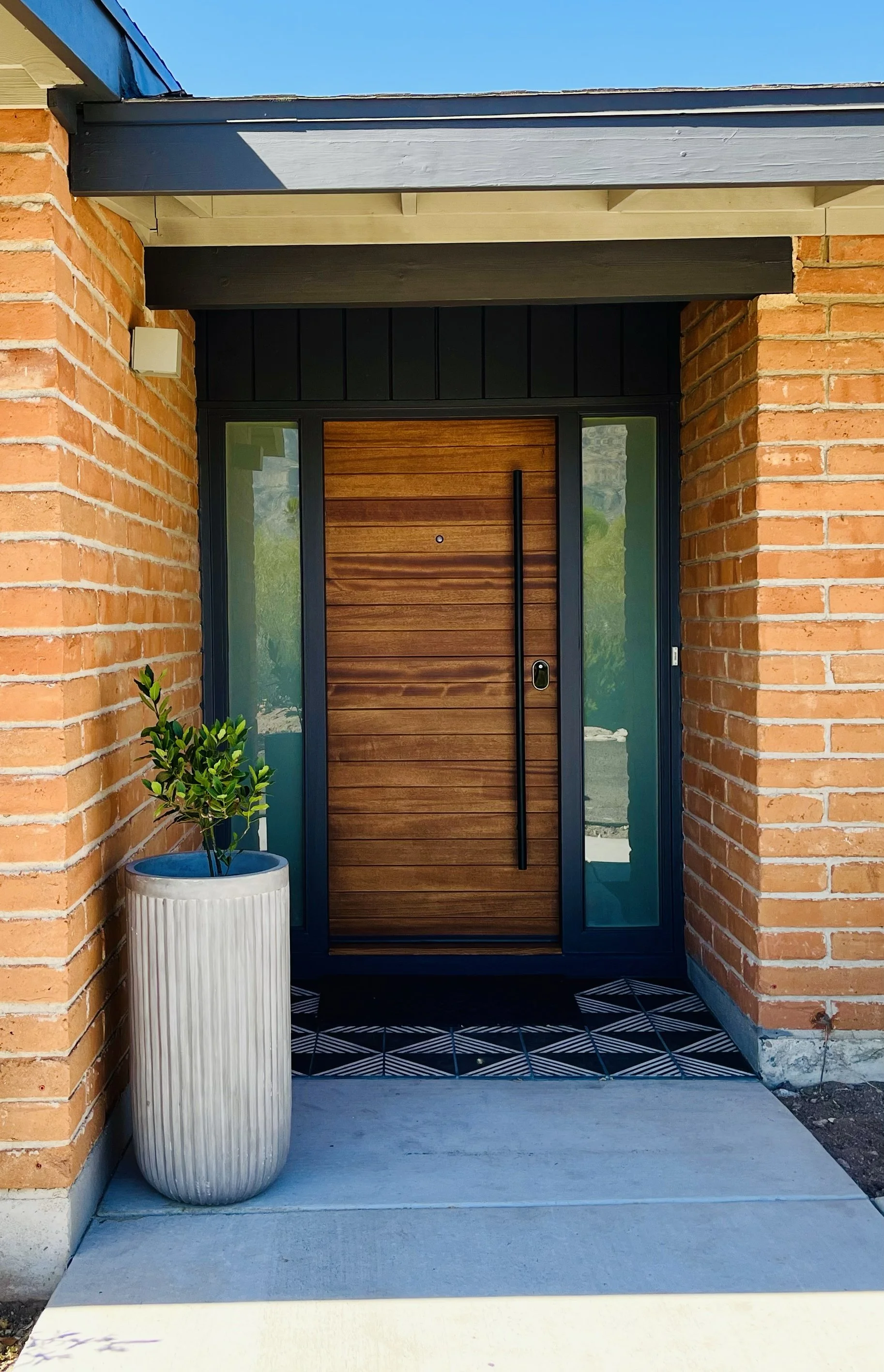 Modern house entrance with wooden door, side glass panels, brick walls, a potted plant, and patterned tile flooring.