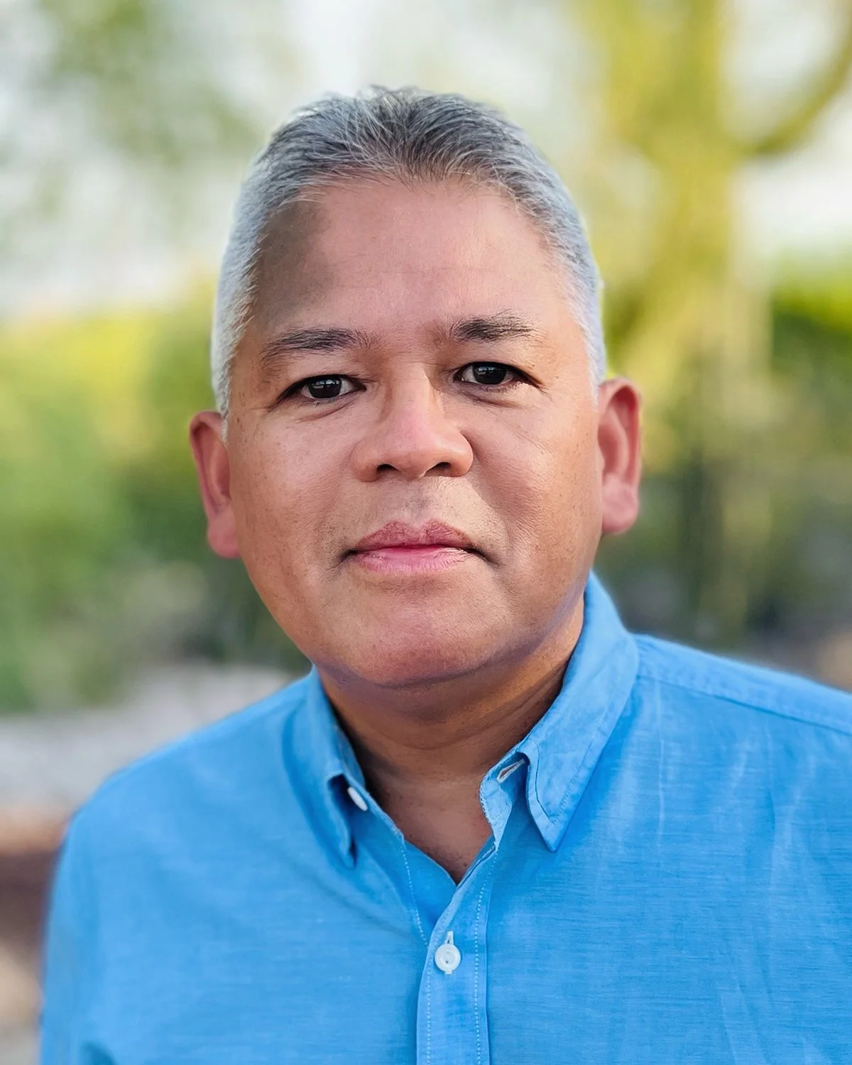Headshot of a middle-aged man with short gray hair, wearing a light blue button-up shirt, outdoors with blurred green and yellow foliage in the background.