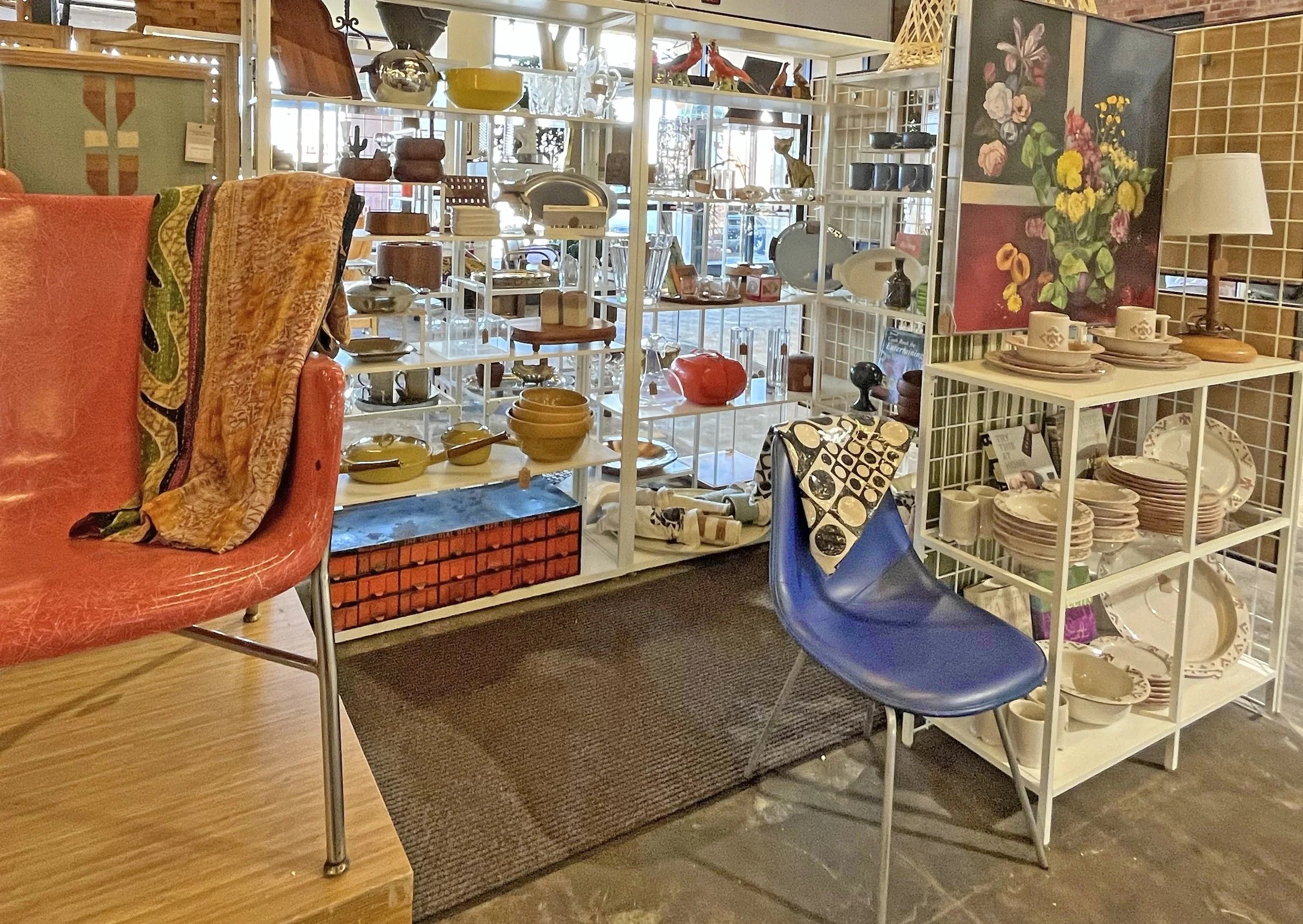 Interior of a vintage home decor store with shelves of ceramics, pottery, and decorative items, including bowls, vases, and figurines, along with a blue chair, a red chair, and artwork on the walls.