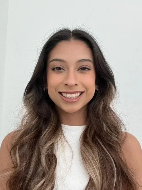 A woman with long wavy brown hair and light skin, smiling, wearing a sleeveless white top, standing against a plain white background.