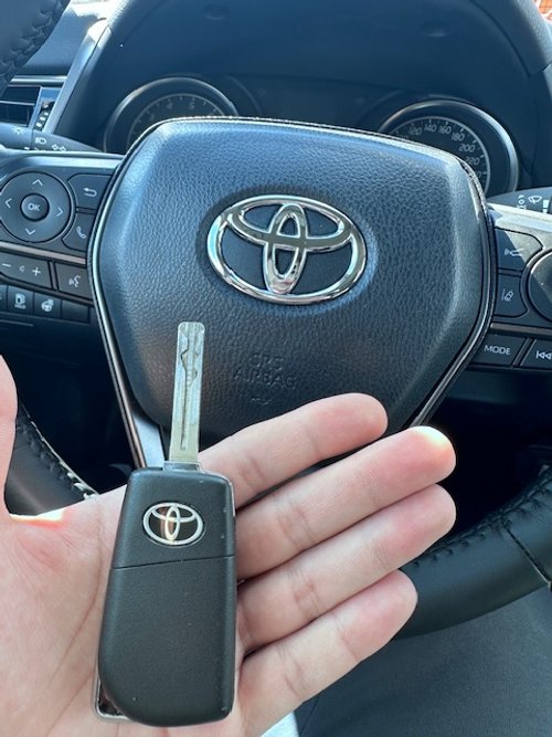 A person's hand holding a Toyota car key in front of a Toyota steering wheel inside a vehicle.