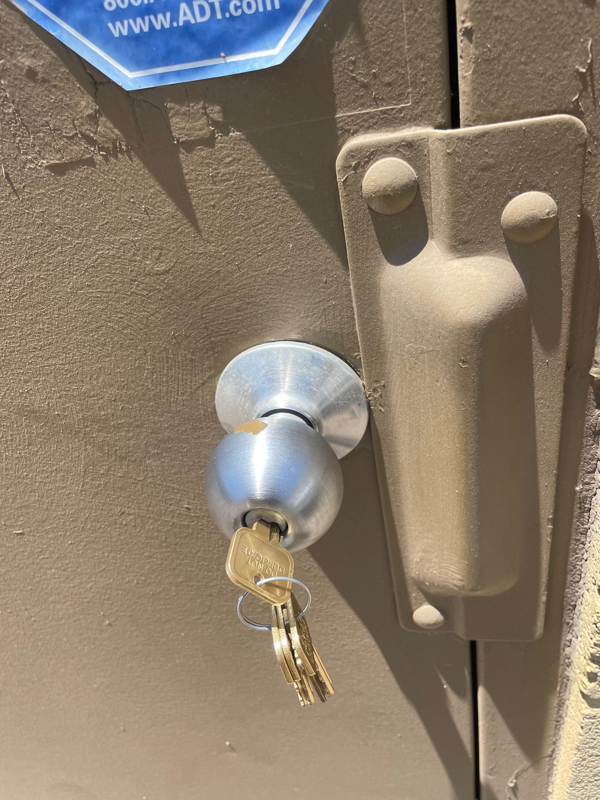 Close-up of a silver door knob with keys inserted, mounted on a door with a metal latch and a textured surface. Part of a blue ADT security sign is visible at the top.