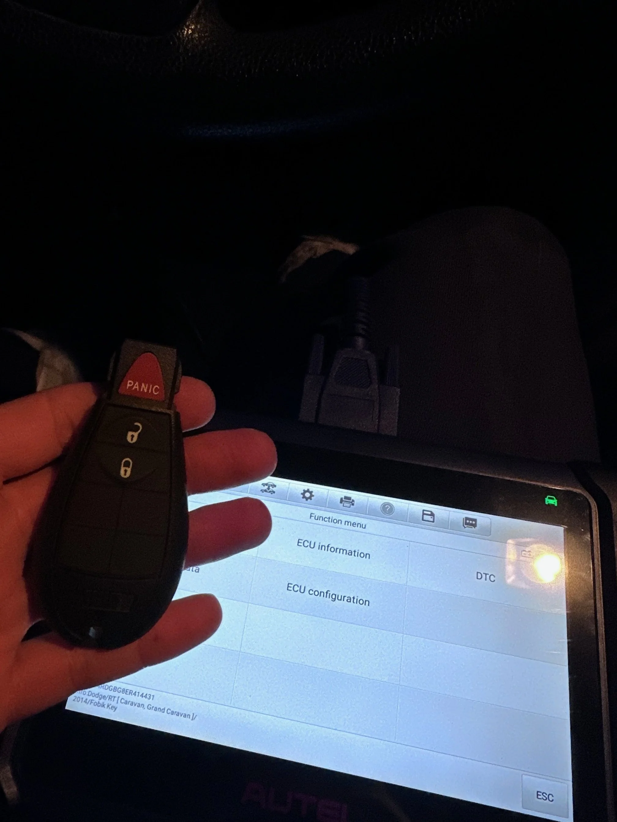 A person holding a car key fob with a red panic button in front of a diagnostic scanner screen inside a vehicle. The scanner screen displays a menu with options including 'Function menu,' 'ECU information,' and 'ECU configuration.'