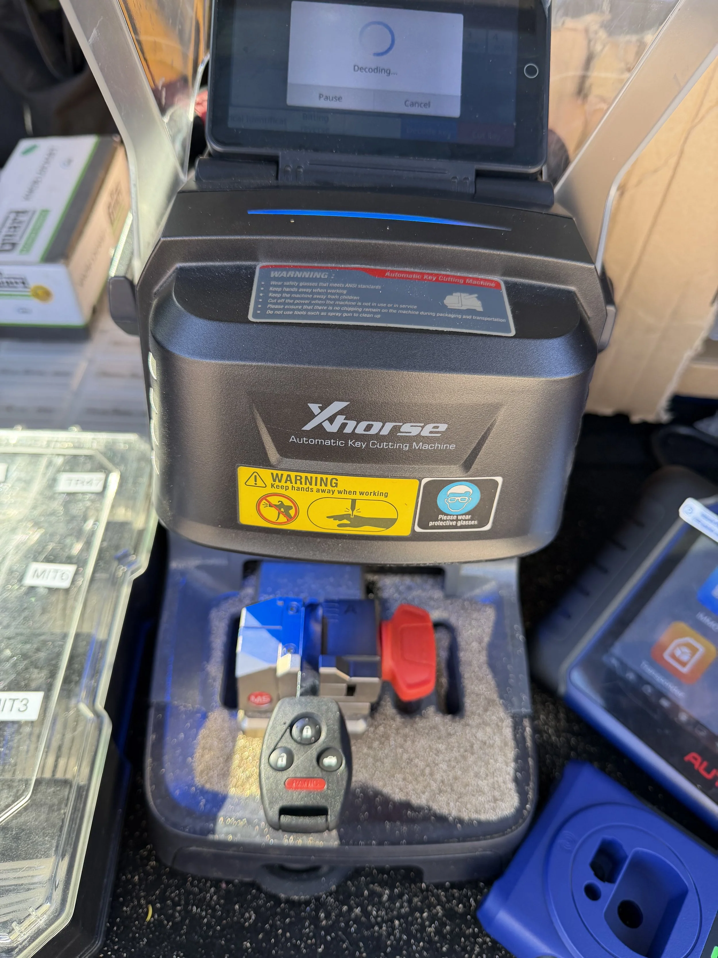 Close-up of an Xhorse automatic key cutting machine with a black and red control unit, warning labels, and a remote control, placed on a black surface with plastic containers and boxes around.