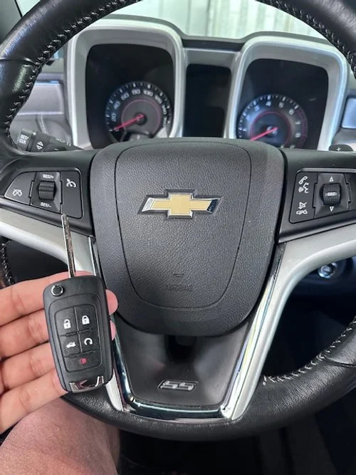 Close-up of a person's hand holding a Chevrolet car key fob with buttons for lock, unlock, and trunk, in front of a Chevrolet steering wheel and dashboard inside a vehicle.