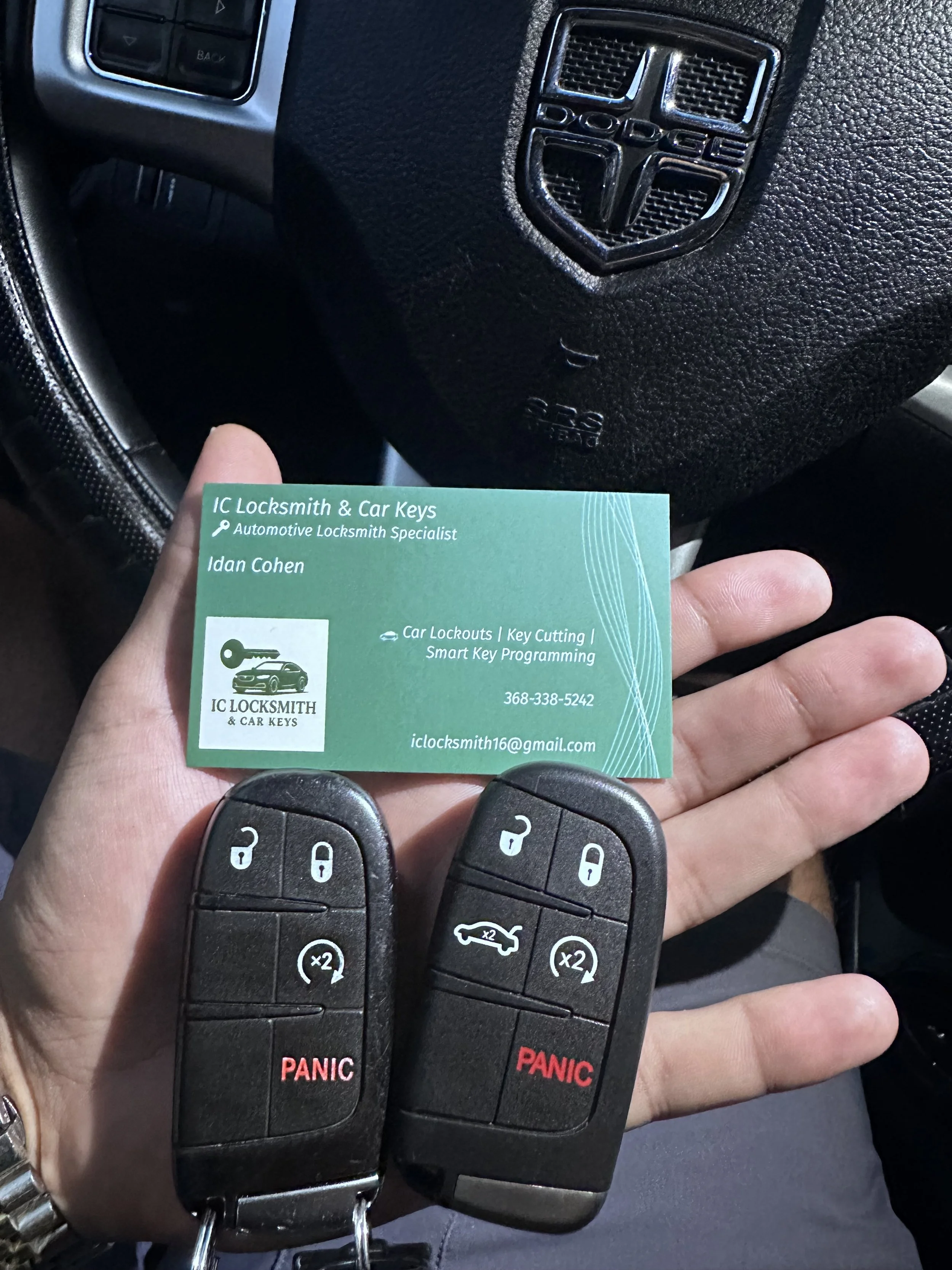 A hand holding two black car remote keys and a business card for IC Locksmith & Car Keys in front of a car steering wheel.