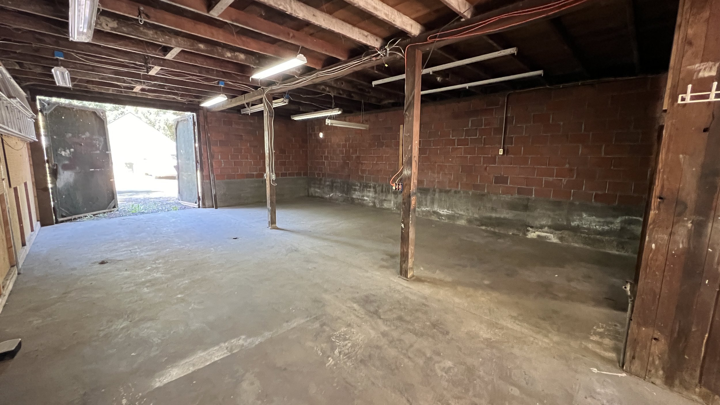 Empty garage with brick wall, wooden beams, exposed wiring, and a concrete floor, with open garage door letting in outside light.