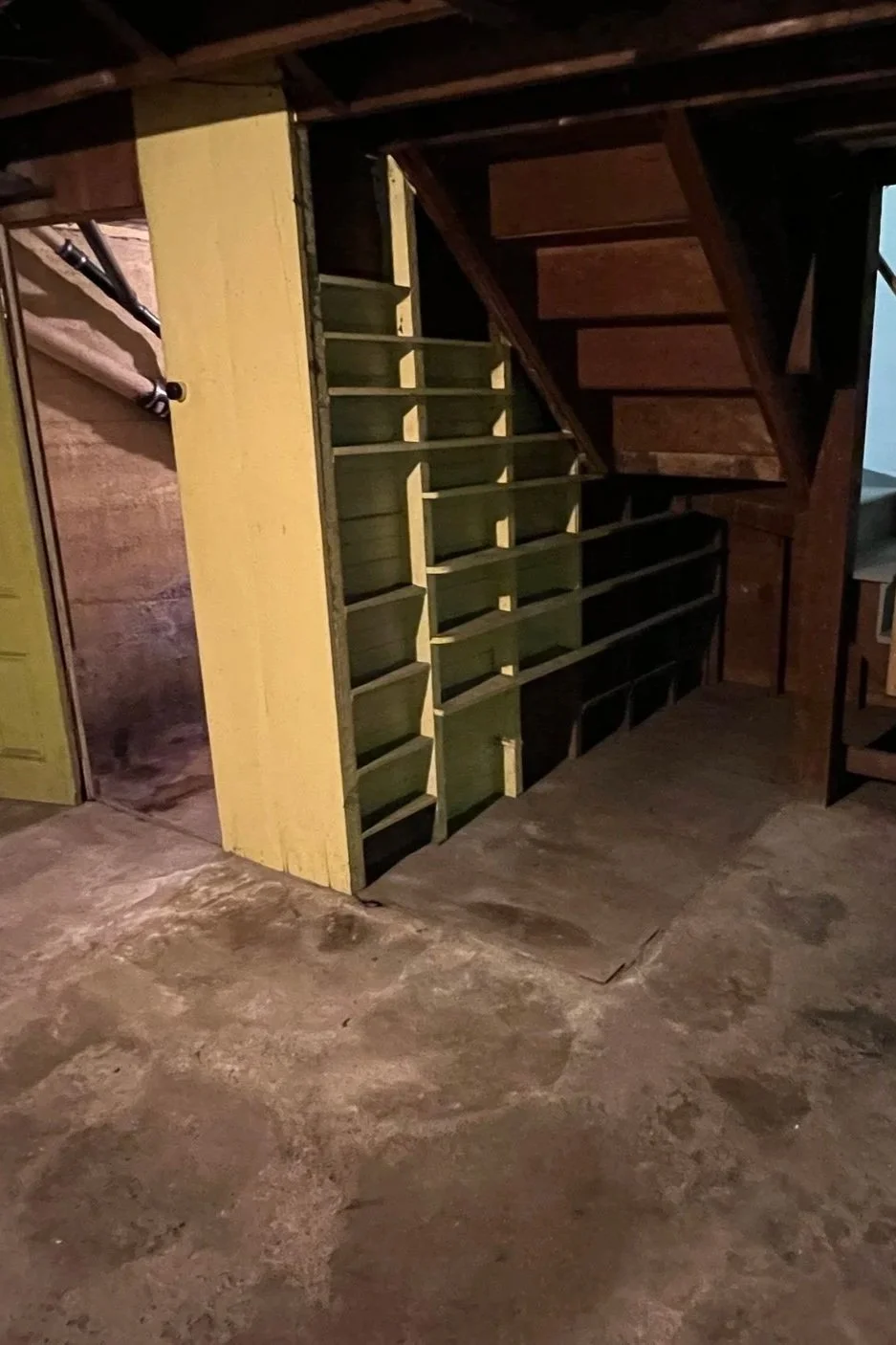 Interior of a basement or cellar with unfinished concrete floor and partially built wooden shelves under a staircase. Completely cleaned out basement.