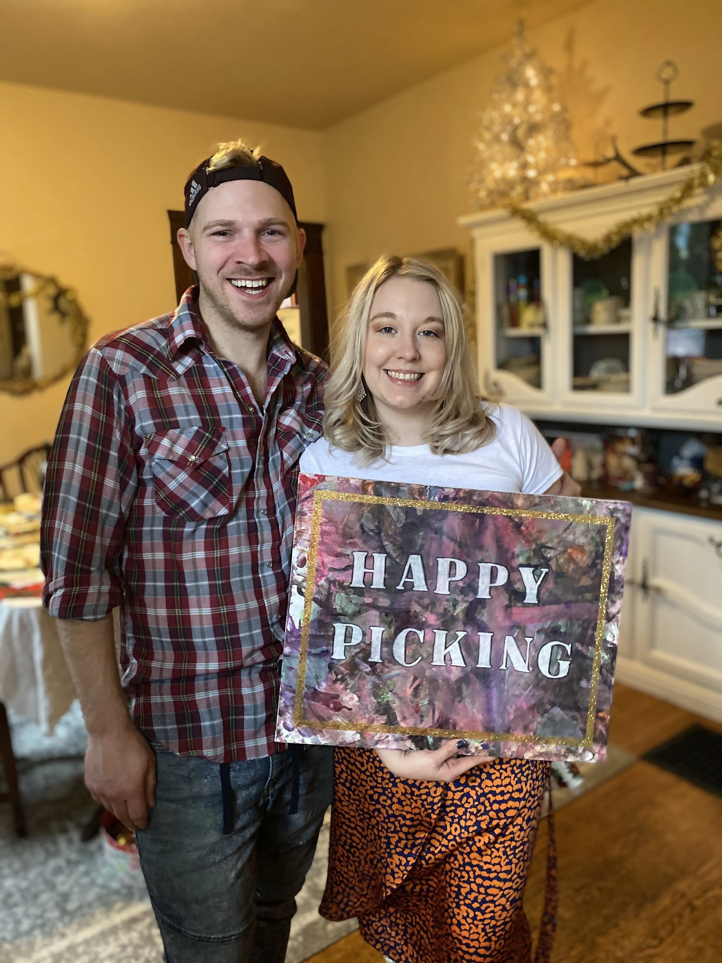 A man and woman holding a sign that says 'Happy Picking'. The man is wearing a backward baseball cap and plaid shirt, and the woman is wearing a white shirt and orange animal print skirt. They are smiling and standing in a decorated living room.
