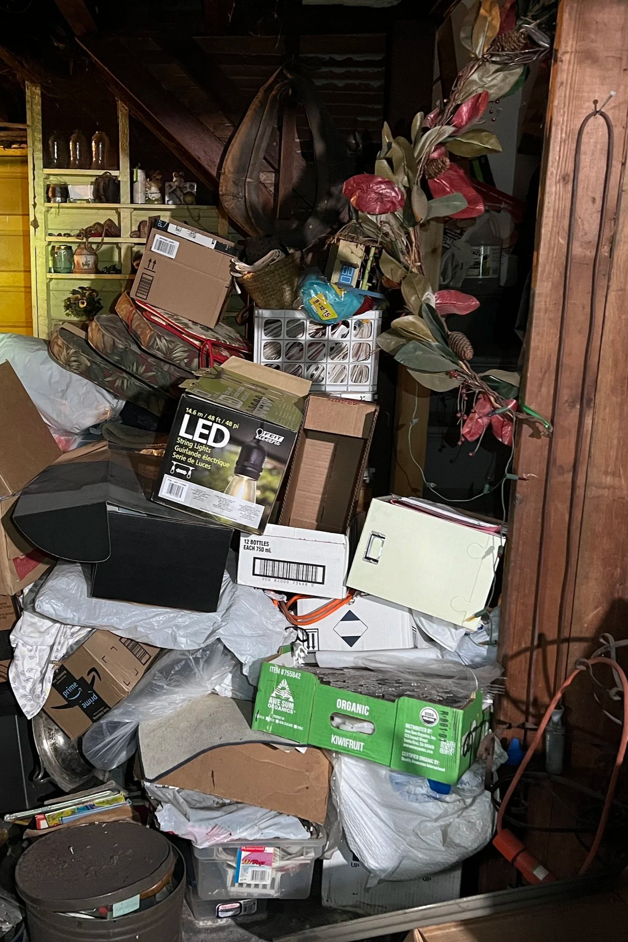 A cluttered storage area filled with boxes, bags, and various household items. There are cardboard boxes, plastic bags, a green organic kiwifruit box, a black LED string light box, and other miscellaneous objects stacked on a shelf and on the floor.