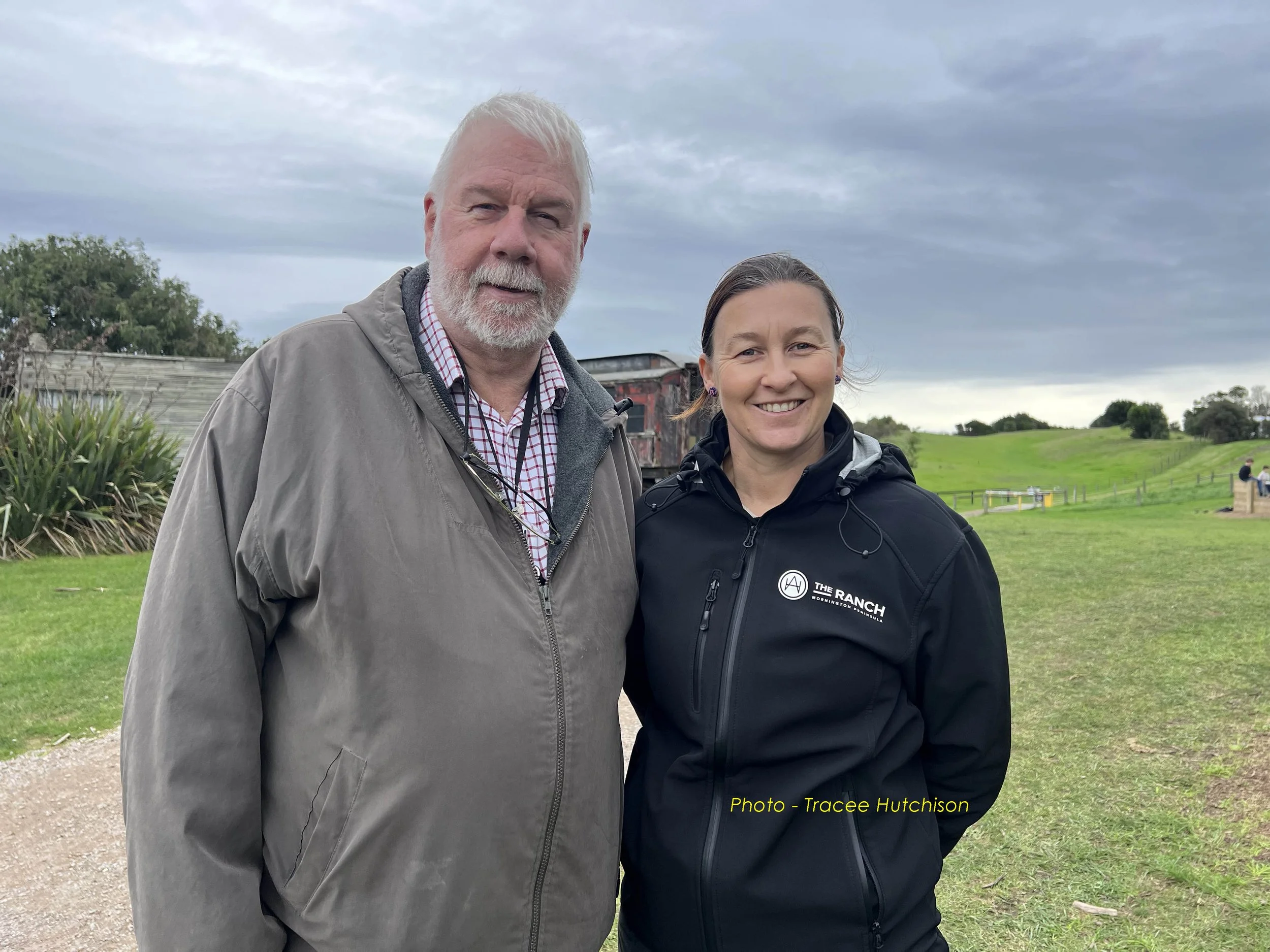A man and a woman standing outdoors on cloudy day, smiling, with a grassy field and trees in the background.