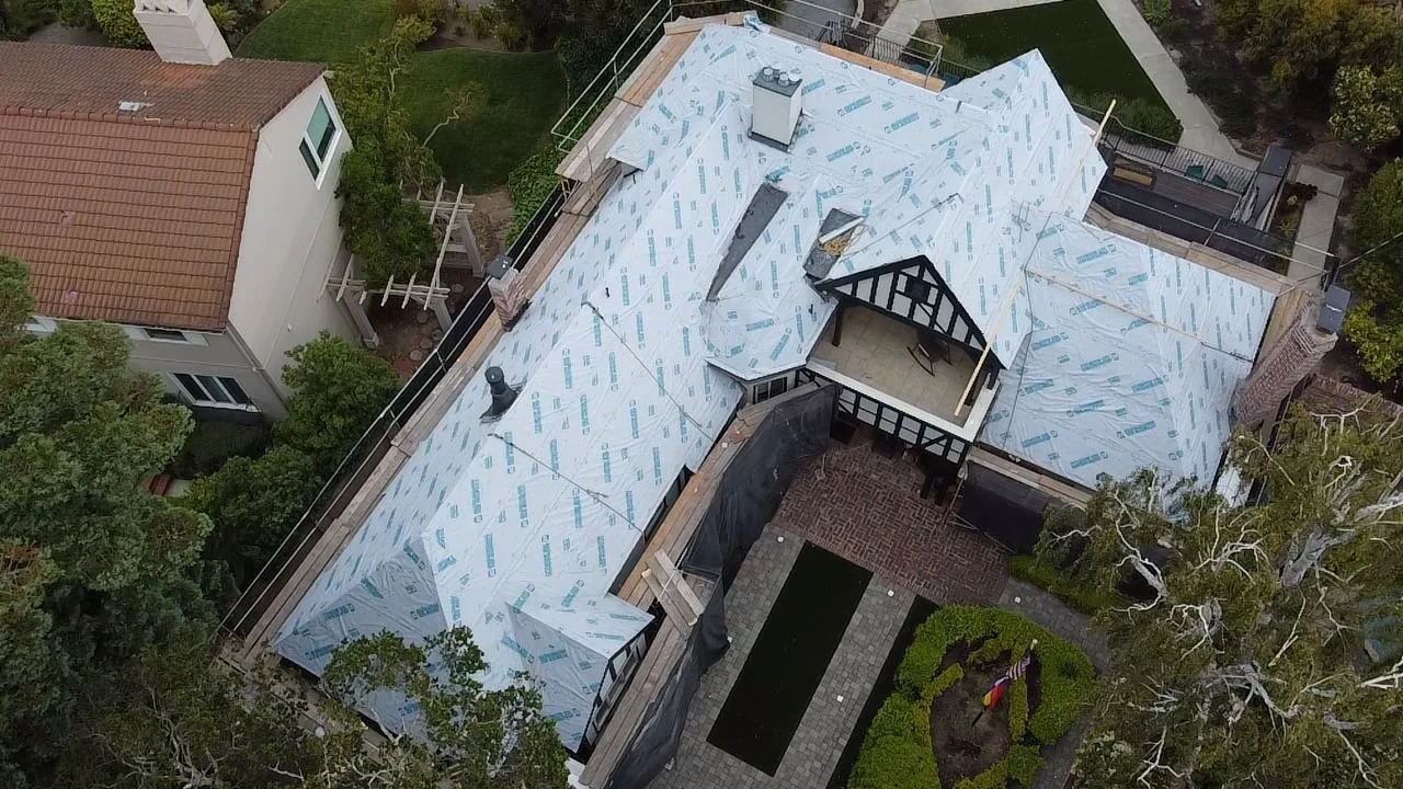 Aerial view of a house under construction or renovation, with a new roof being installed, surrounded by trees and neighboring buildings.