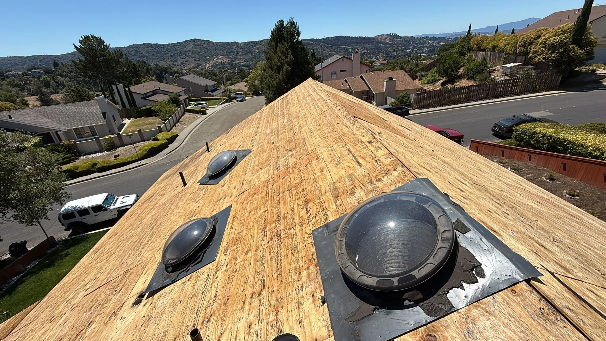 View of a rooftop with new wooden sheathing and ventilation pipes, overlooking a residential neighborhood with parked cars and hilly landscape in the background.