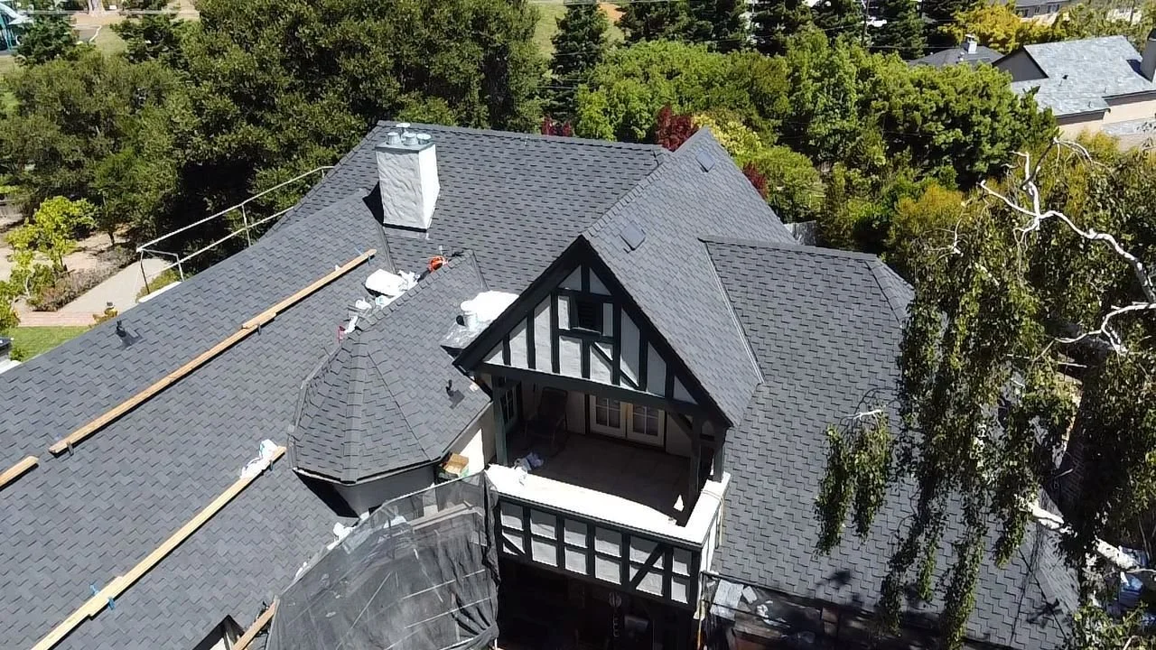 Aerial view of a house with a steep gray shingle roof, a balcony with a balcony railing, and surrounding lush green trees.