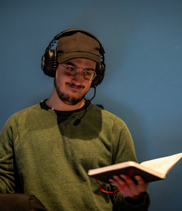 A man with glasses, wearing a green sweater, a brown cap, and a headset, holding an open book and smiling against a blue wall.