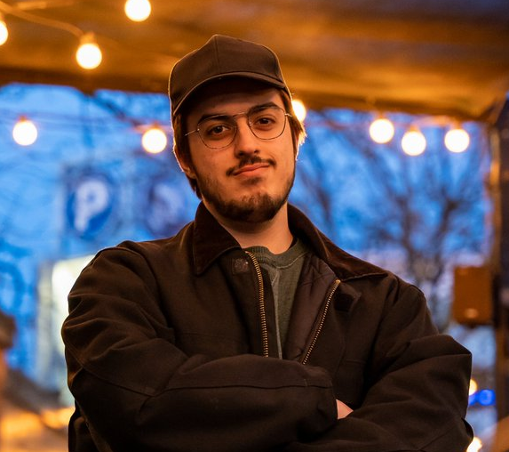 Young man with glasses, beard, and mustache wearing a black jacket and black cap standing with arms crossed in an outdoor setting at dusk, with blurred lights and trees in the background.