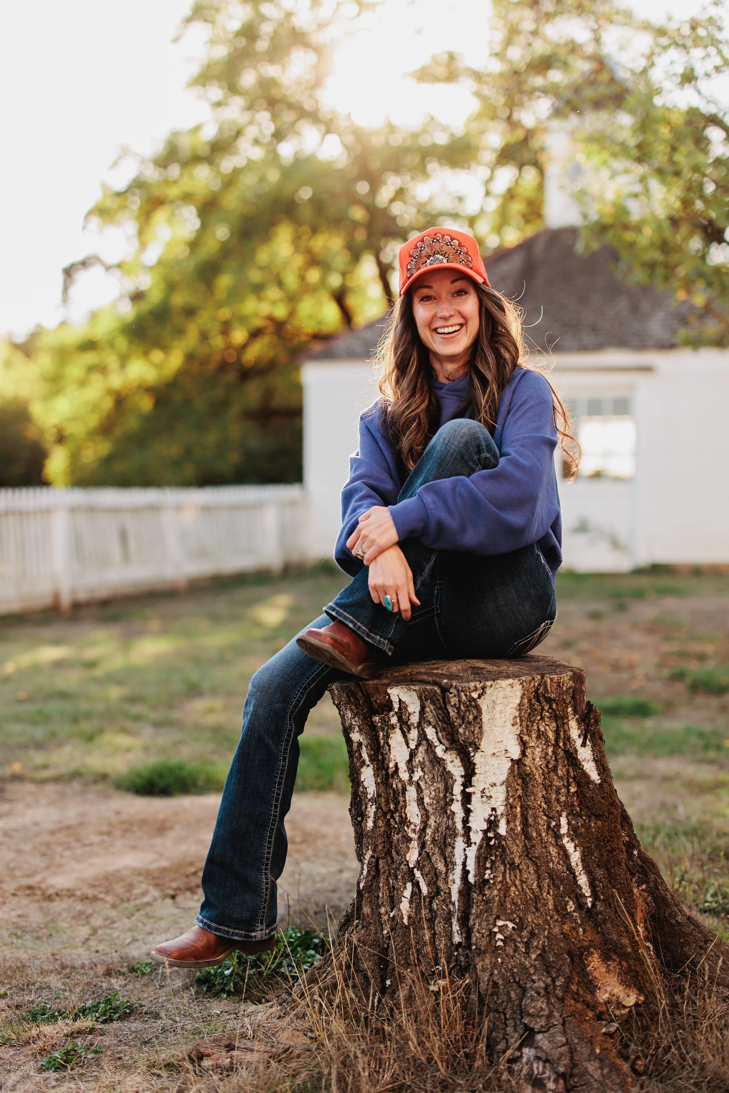 A woman sitting on a tree stump outdoors, smiling, wearing an orange feather crown trucker hat, blue hoodie, dark jeans, and brown boots, with a house and trees in the background during daytime.