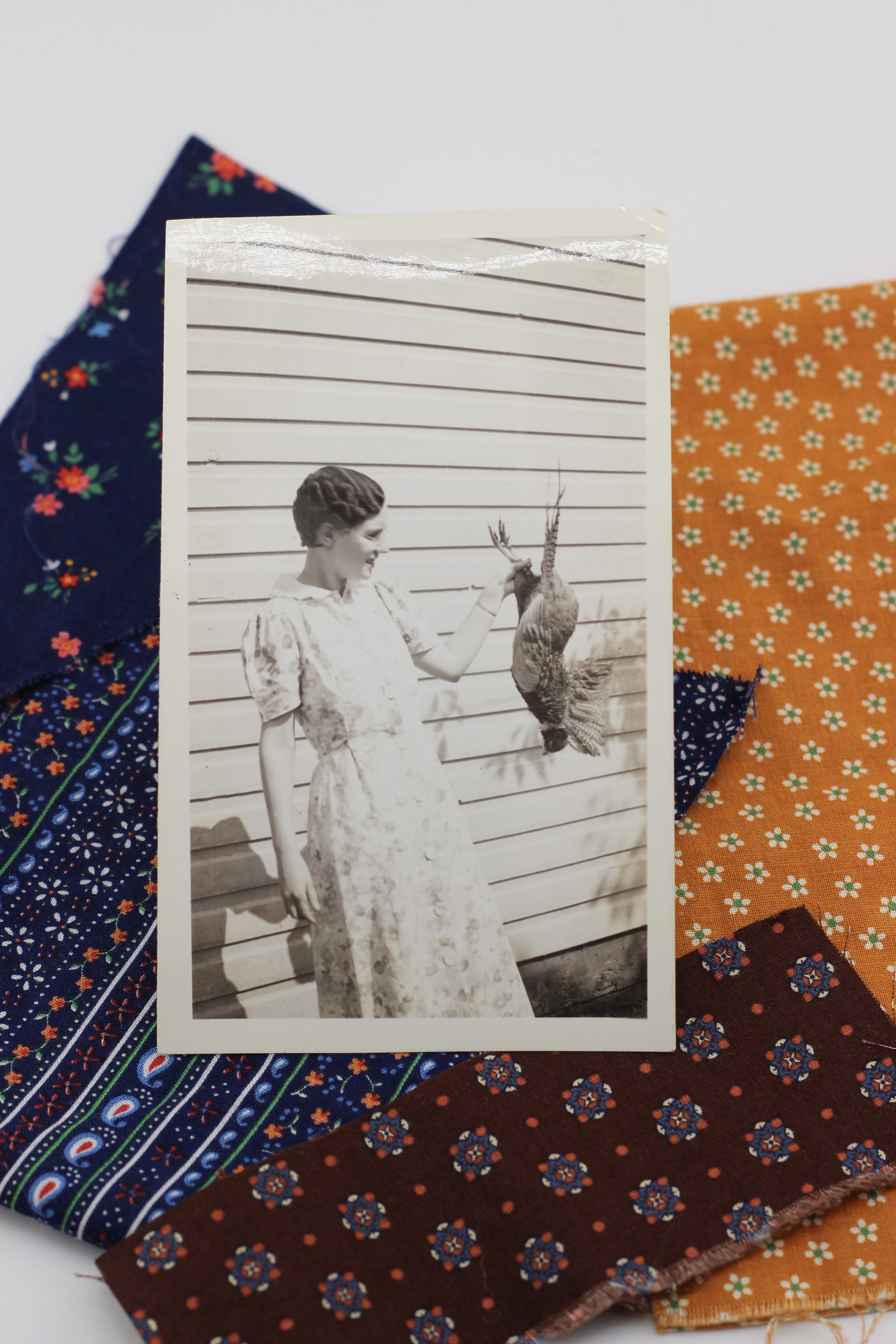 Black and white photograph of a woman in a patterned dress holding a ring neck pheasant rooster she harvested standing against her 1930's house.
