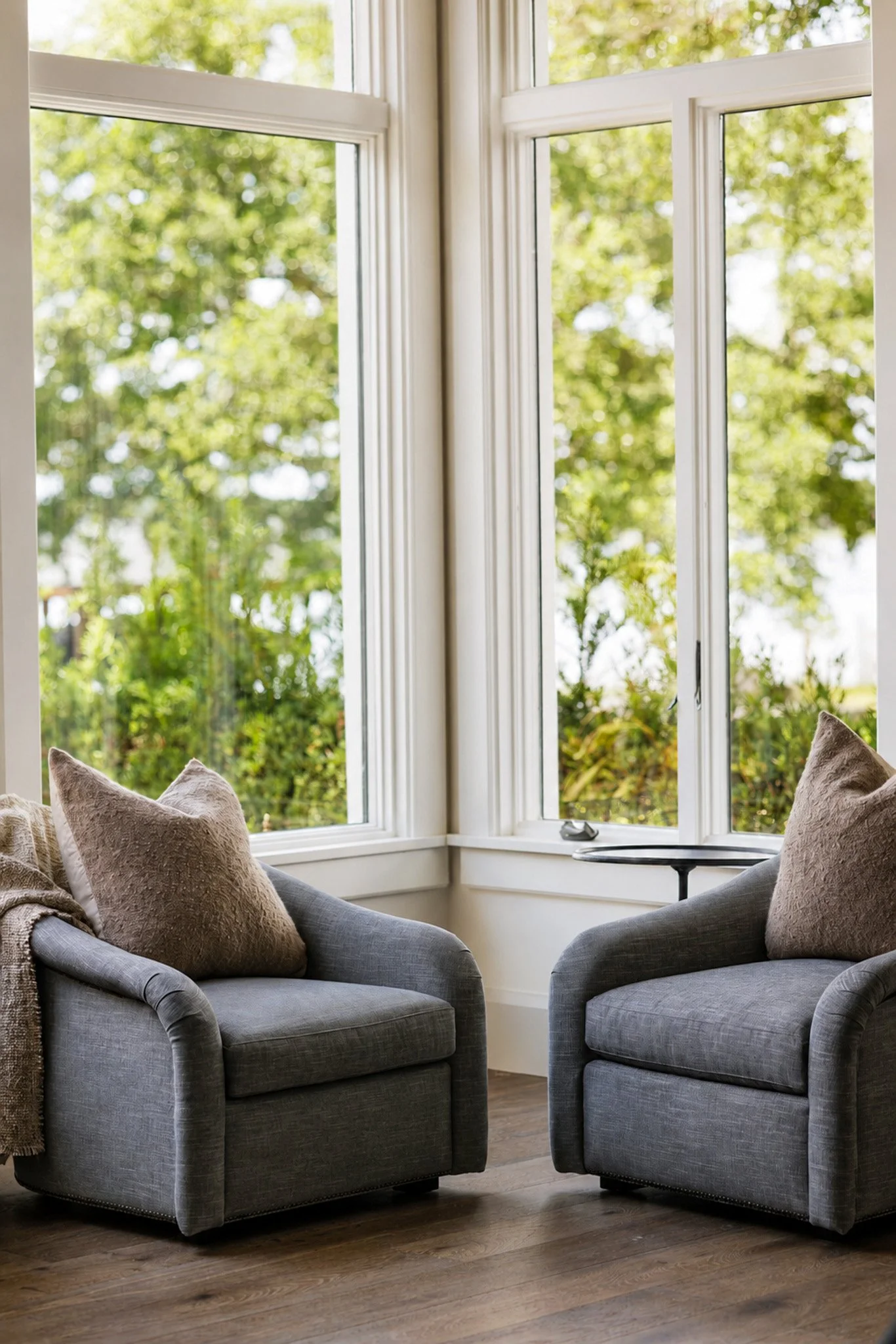 Two gray armchairs with beige pillows are positioned around a small black side table, near large windows with a view of green trees outside.