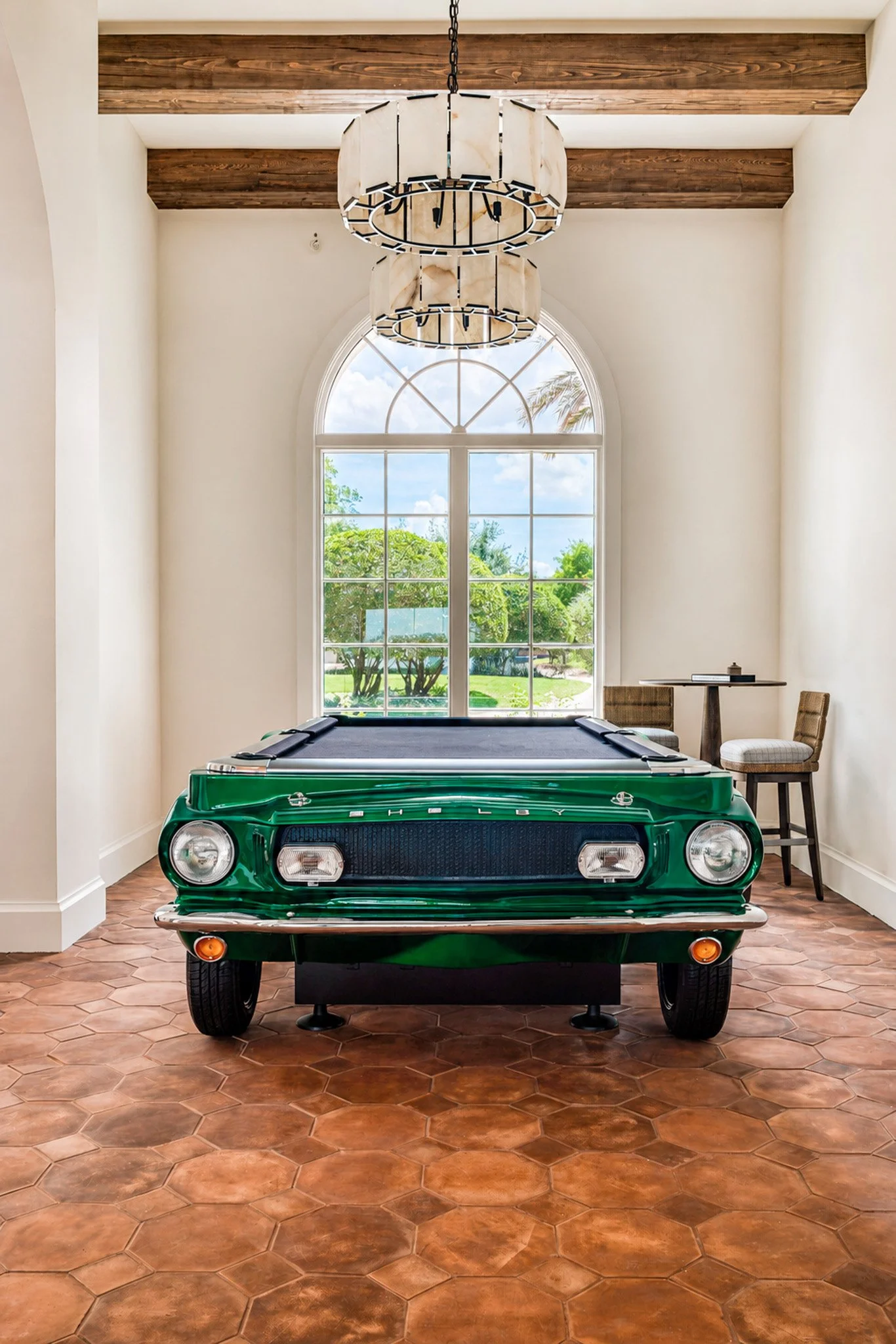 Green vintage car with a black roof placed indoors, with a large arched window, wooden beams on the ceiling, and a chandelier hanging above, with two chairs and a small table to the side.
