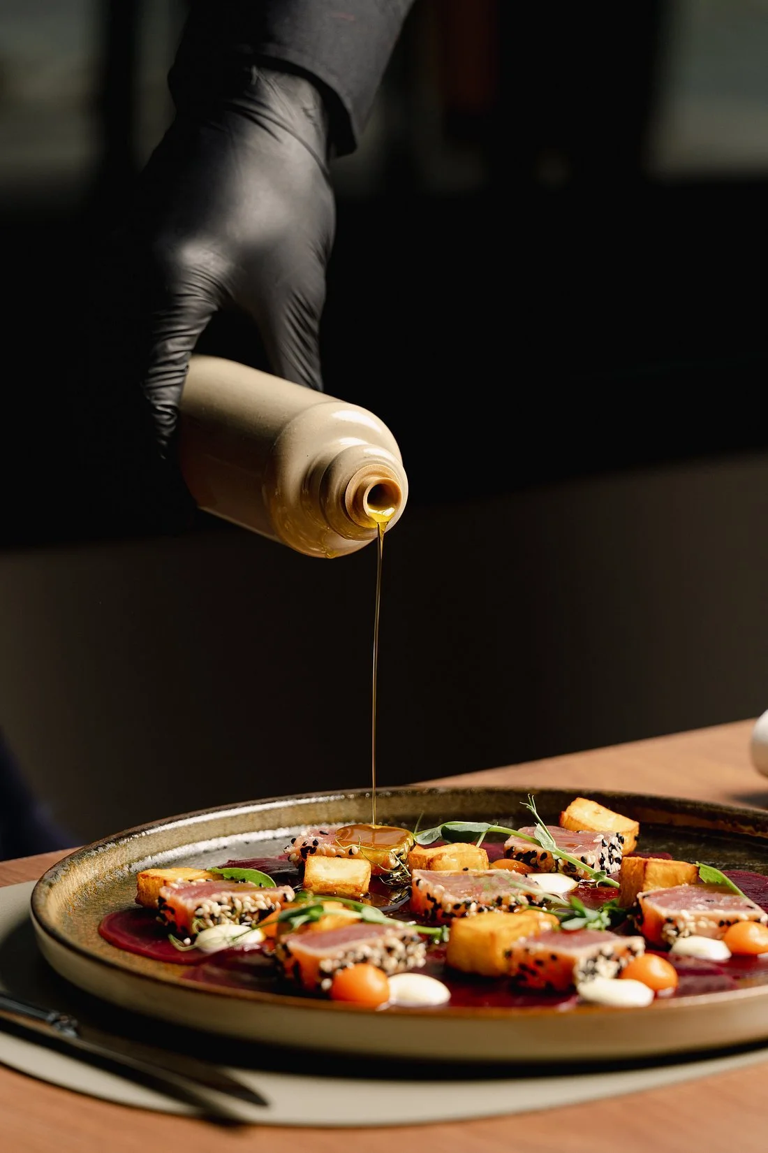 A chef wearing black gloves is pouring sauce over a plated dish of seared tuna with sesame seeds, garnished with greens, dollops of sauce, and vegetables, on a round ceramic plate.