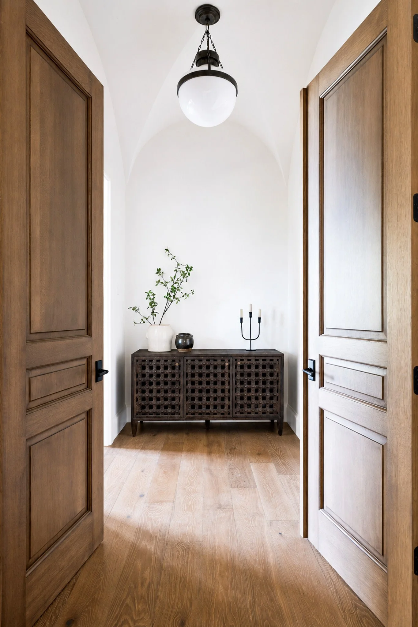 View of a hallway with wooden double doors open, revealing a white wall with a black console table decorated with vases, a potted plant, and candles, topped with a simple black chandelier.