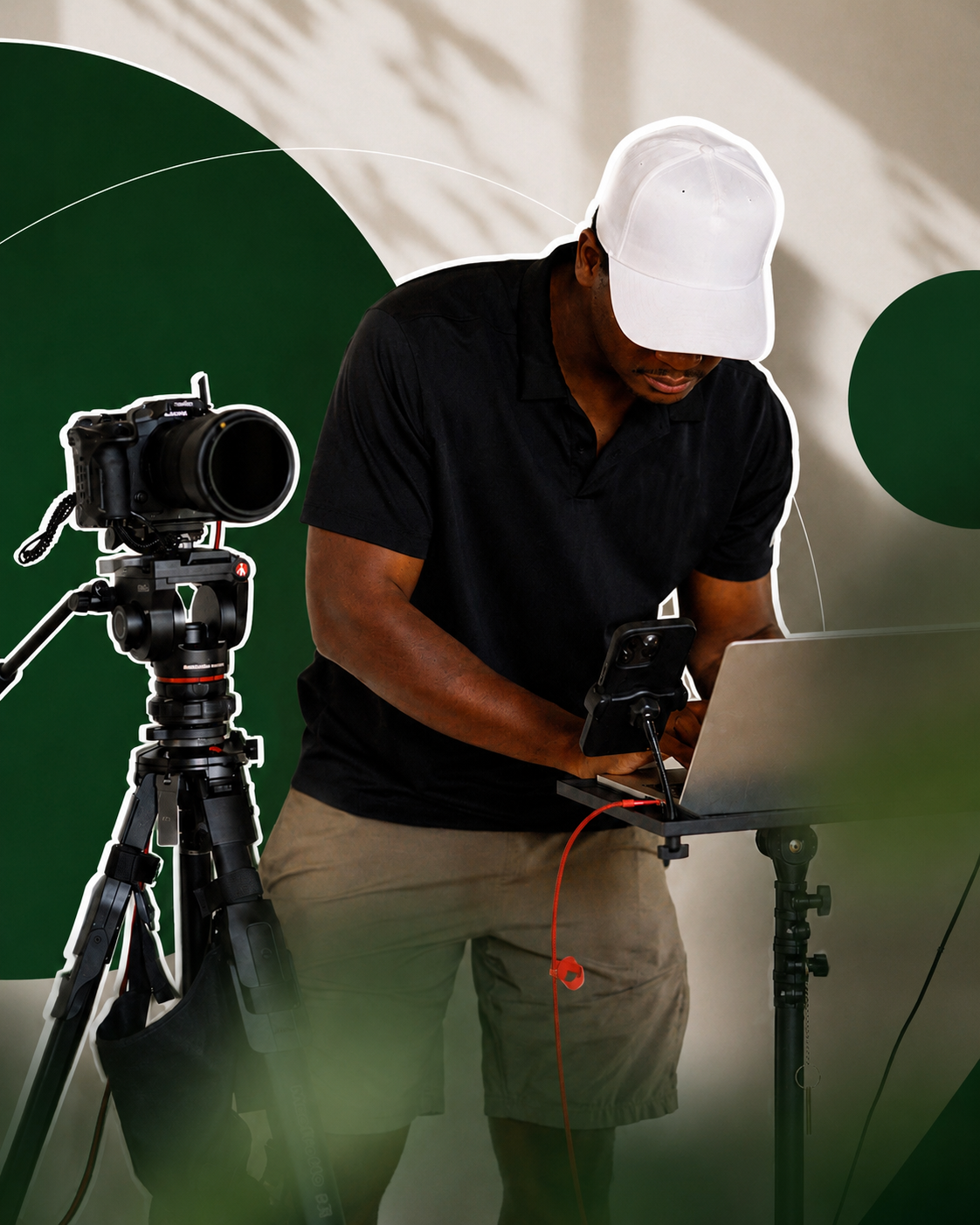A man wearing a white cap, black shirt, and khaki shorts working on a laptop at a desk with a professional camera on a tripod nearby, against a green and white background.