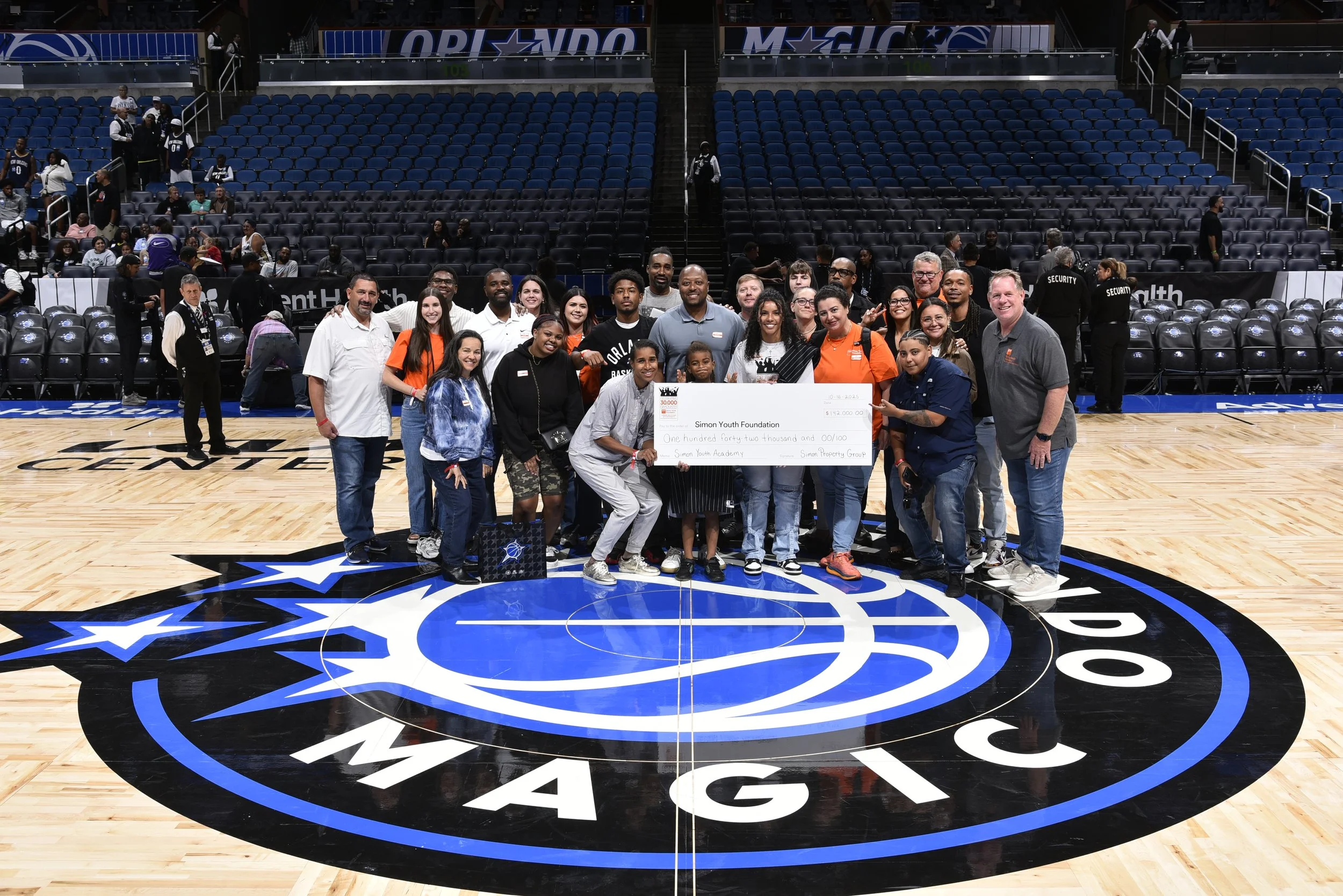 Group of diverse people, including children, teens, and adults, standing on a basketball court at Amway Center, holding a large check from the Simon Youth Foundation. The background shows empty seats and some security personnel.