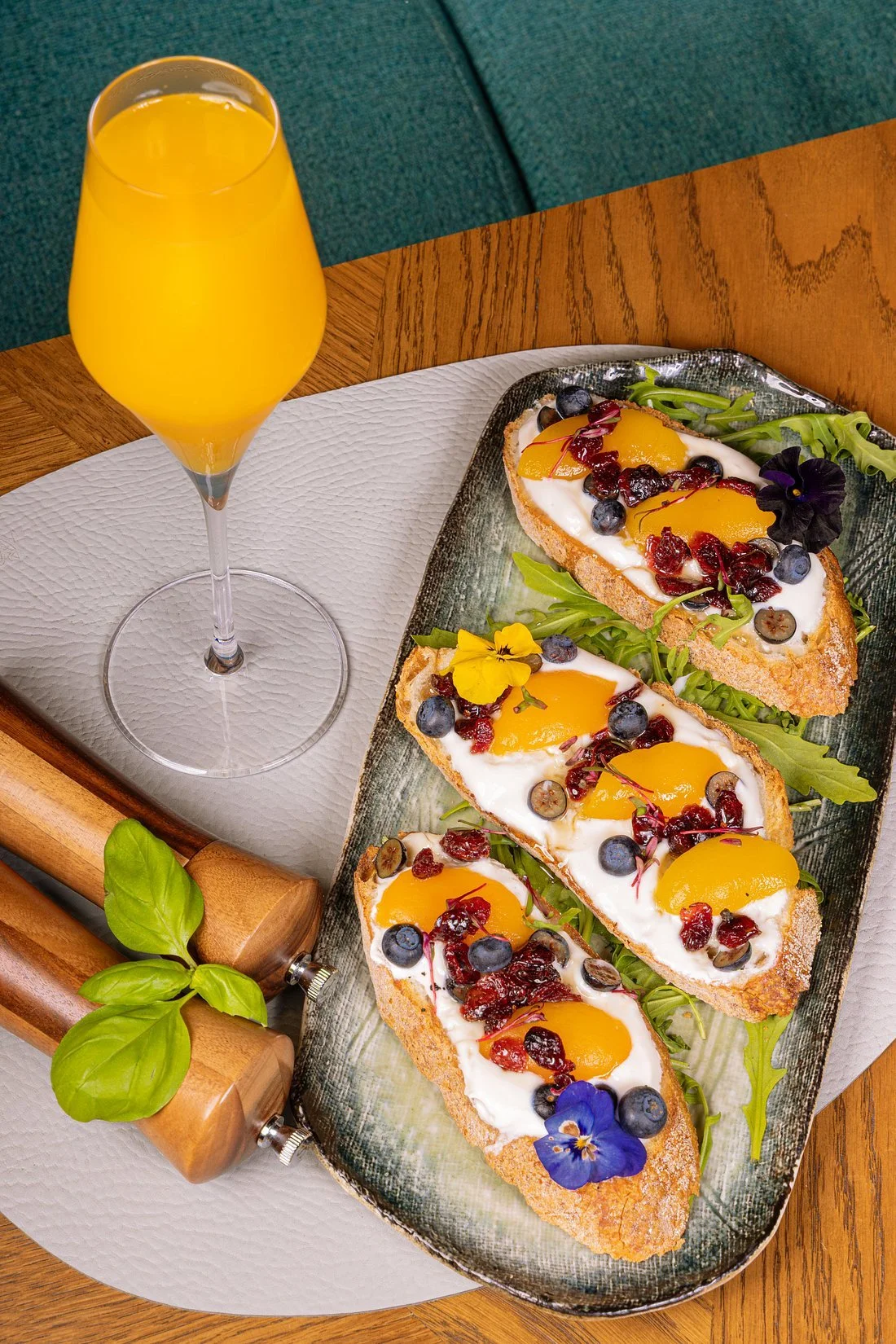 A glass of orange juice, three pieces of avocado toast topped with eggs, blueberries, dried cranberries, edible flowers, and greens, on a ceramic serving dish on a wooden table.