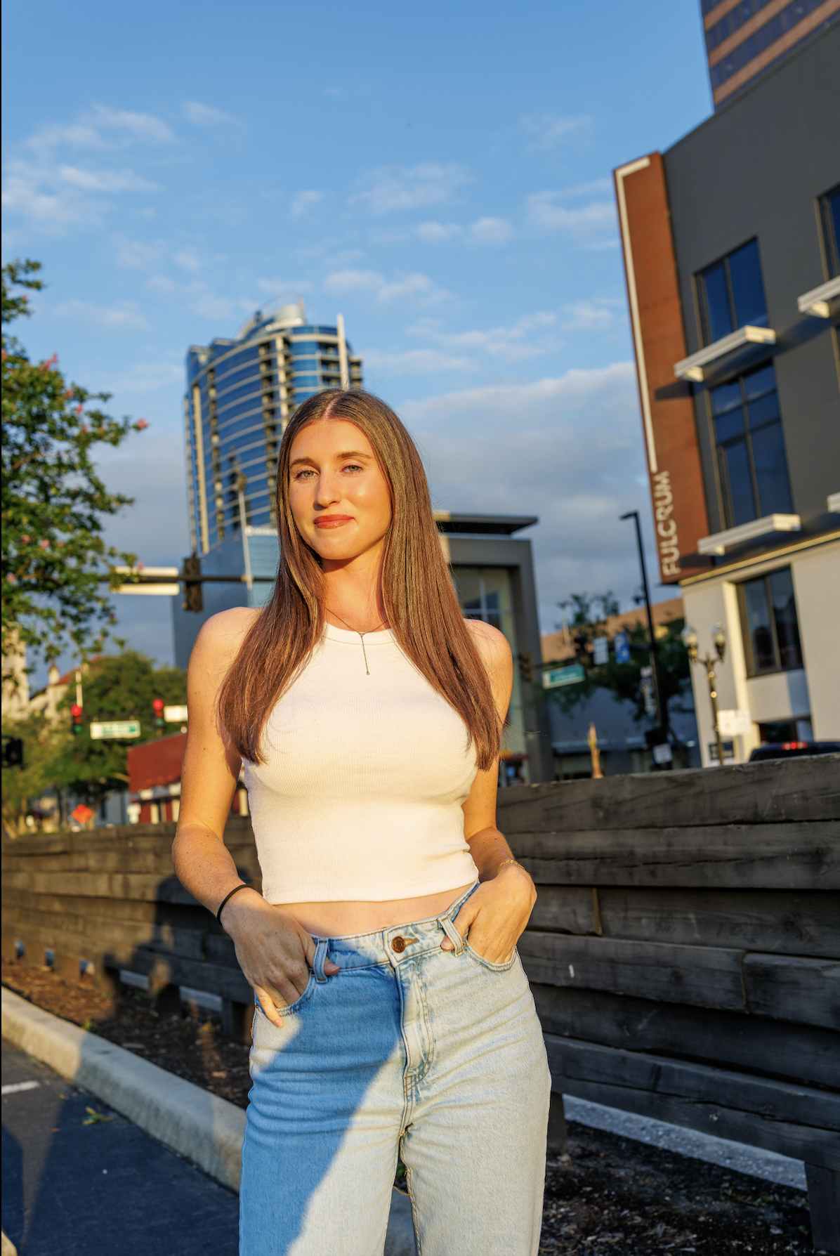 A young woman with long brown hair standing outdoors on a city street during golden hour, wearing a white sleeveless top and light blue jeans, with modern buildings and blue sky in the background.