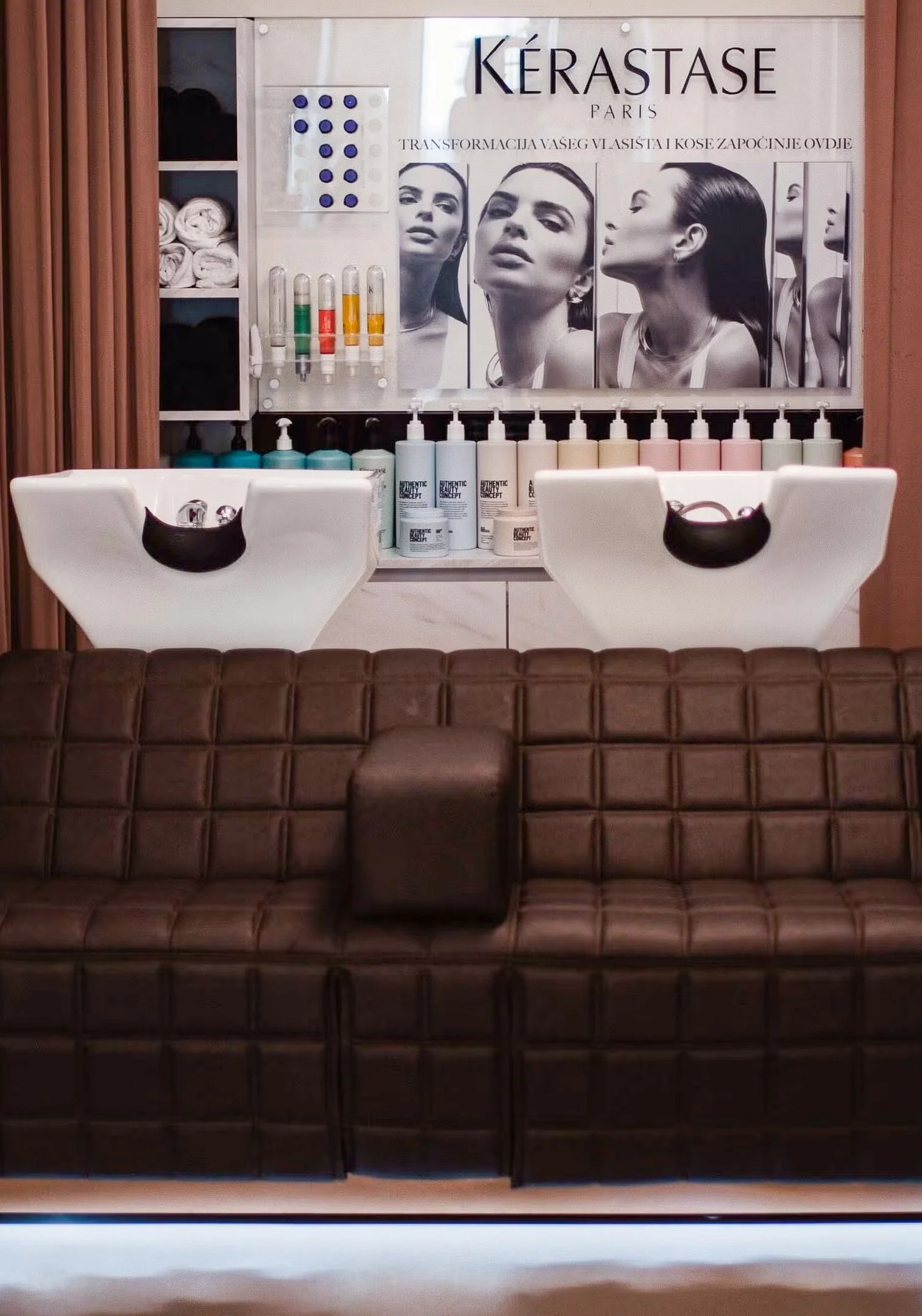 Salon setup with two white hair washing sinks, shelves with hair products, and a large poster featuring black-and-white portraits of women with straight hair. The poster displays the brand name 'Kérastase Paris' and some text in French.